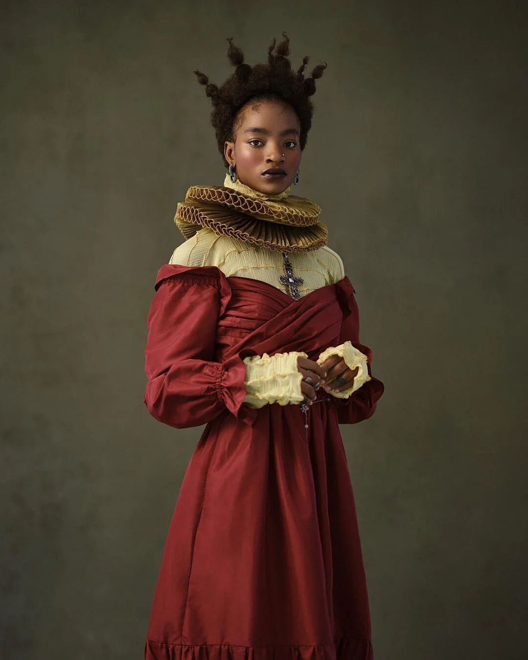A woman with dark skin and styled hair wearing a layered, Victorian-inspired dress with ruffled cuffs, and a large, ruffled collar. She has jewelry including earrings and a necklace with a cross pendant. Her expression is serious, and the background is a plain, dark, muted color.