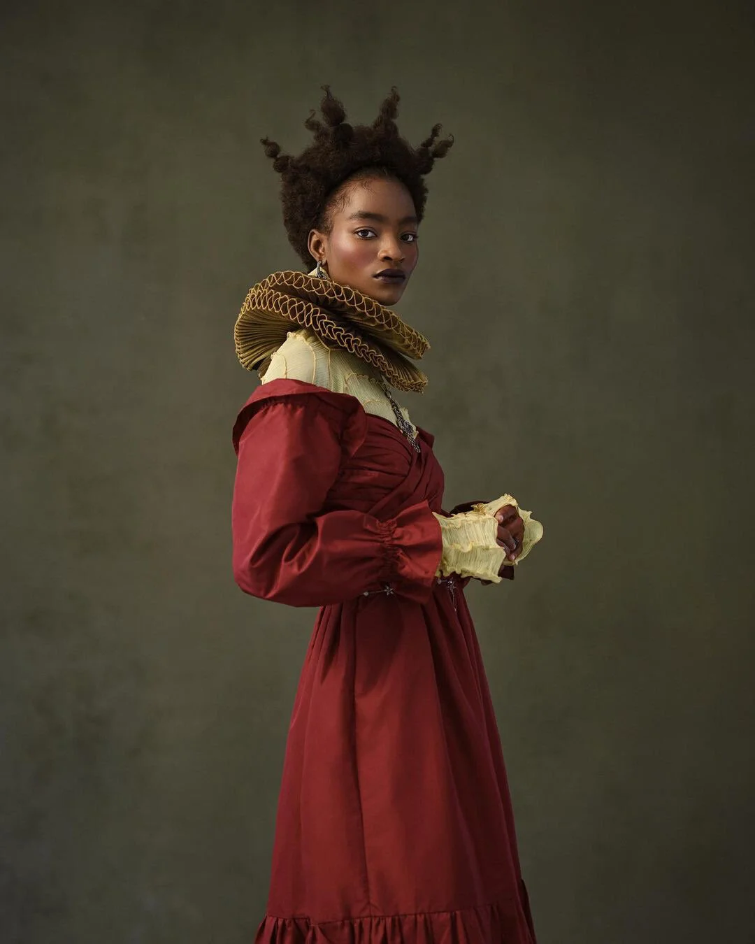 A woman with natural, styled hair wearing an elaborate outfit featuring a red dress with puffed sleeves, a yellow and ruffled high-neck collar, and matching yellow ruffled gloves, posing against a plain background.