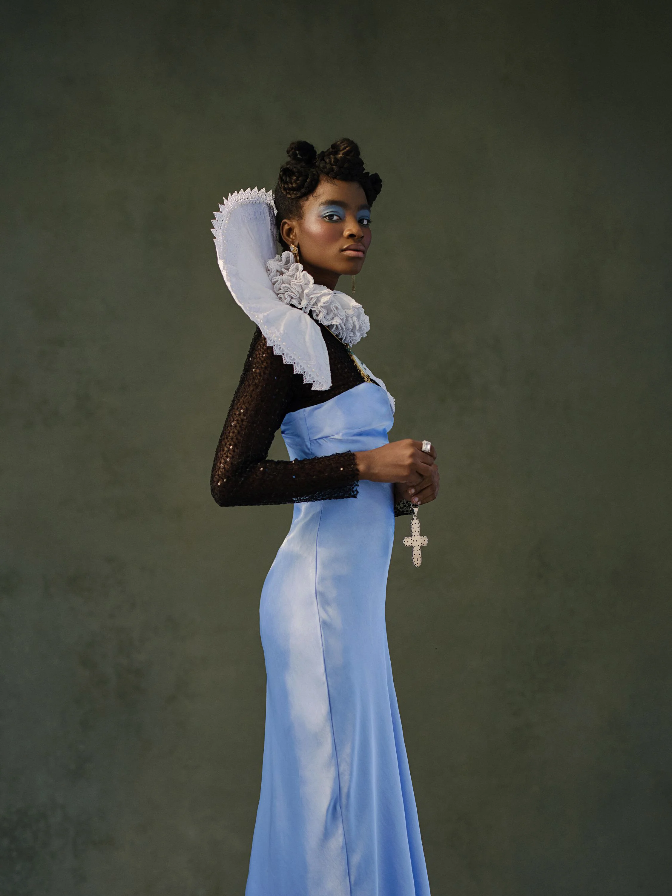 A woman in a blue satin gown with a black sequined long-sleeve top, a large white ruffled collar, and holding a large cross necklace. She has styled hair and blue eye makeup, standing against a plain dark background.