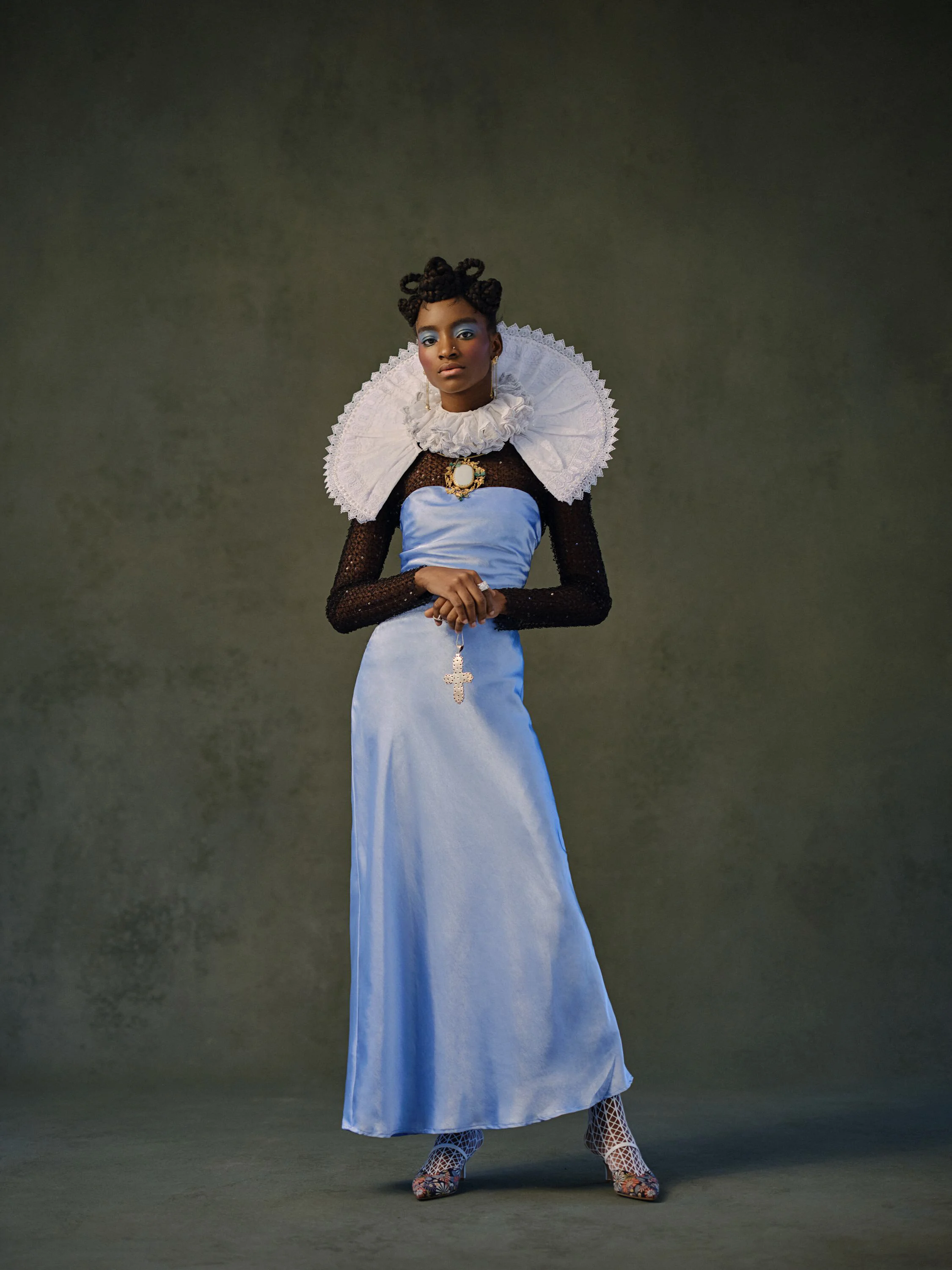 A woman in a high fashion outfit with a black sheer long sleeve top, a blue satin dress, and lace shoes, standing against a dark background with a serious expression.