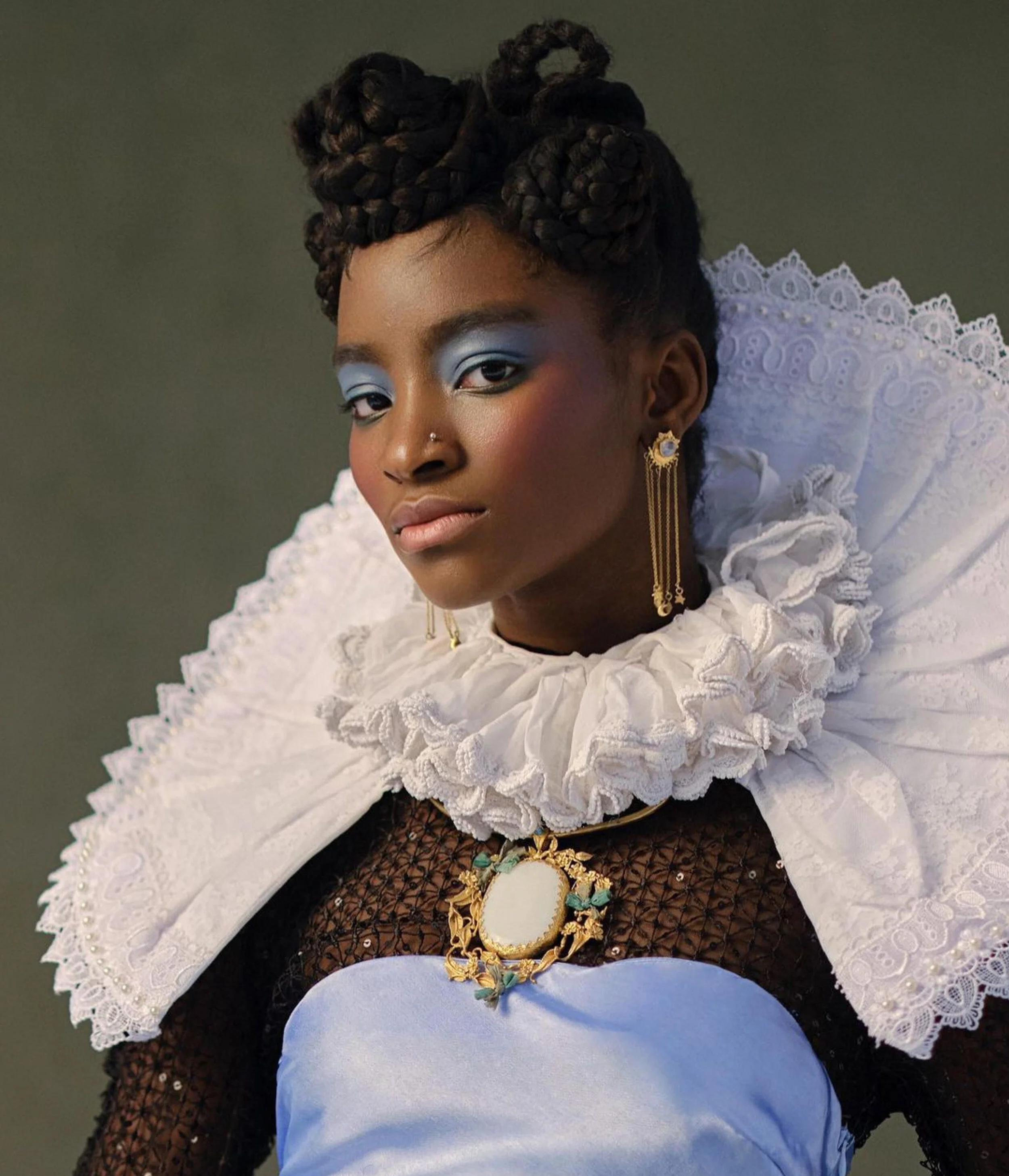 Portrait of a woman with styled braids, wearing a ruffled white collar, black mesh top with sequins, and ornate jewelry, including earrings and a large pendant with a mirror and floral details.