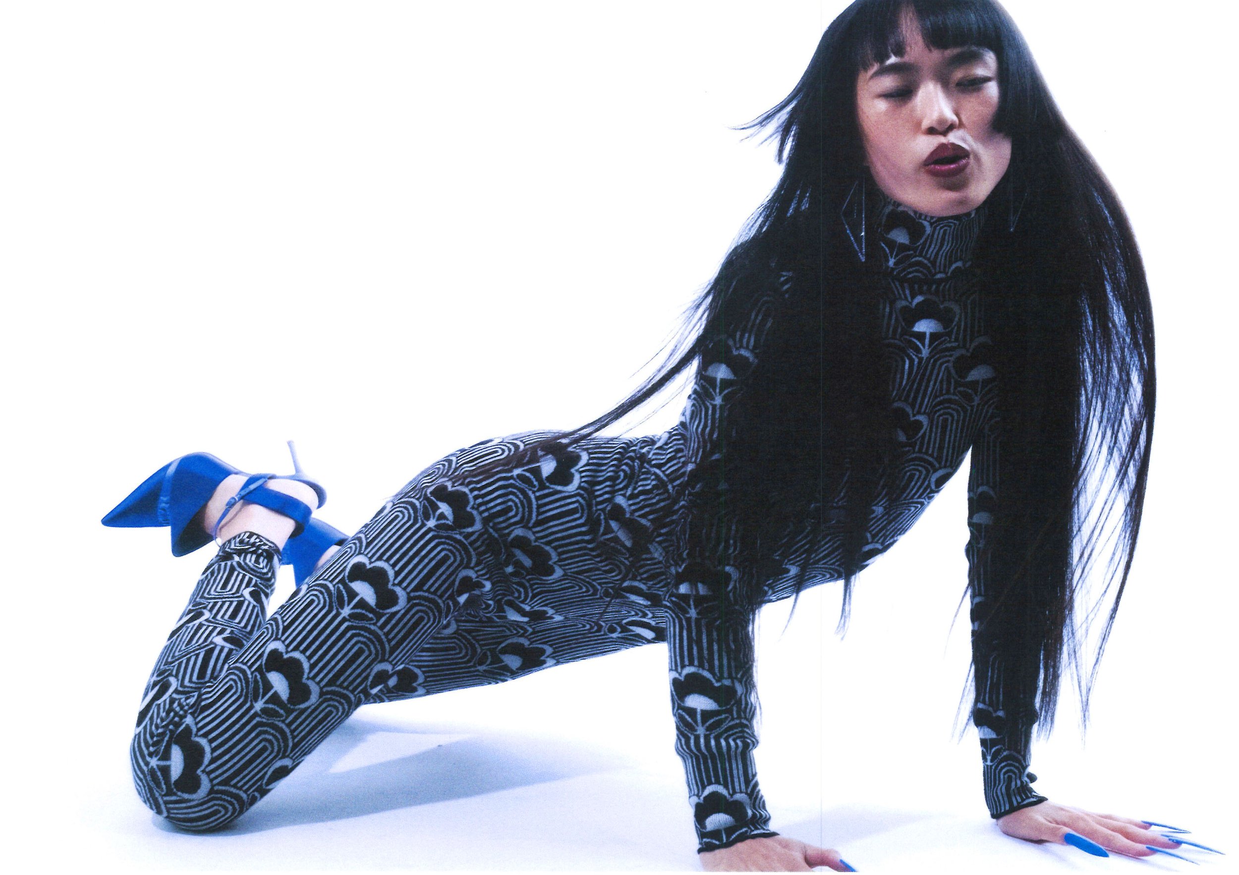 Person with long black hair wearing a patterned black and white outfit, blue high heels, and posing with hands and knees on the ground, against a plain white background.