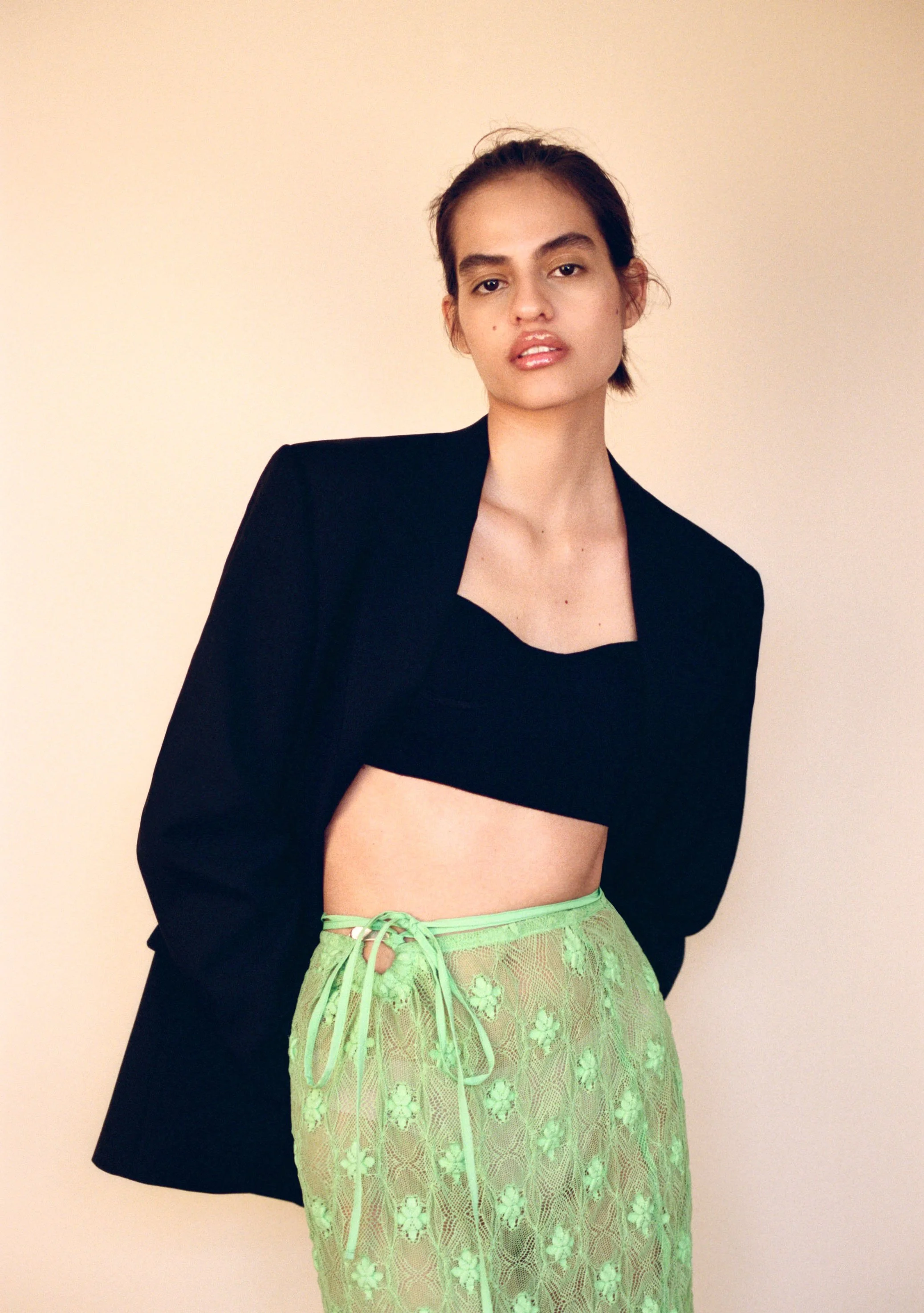 A young woman with brown hair, wearing a black blazer, a black crop top, and neon green lace pants with floral details, standing against a plain beige background.