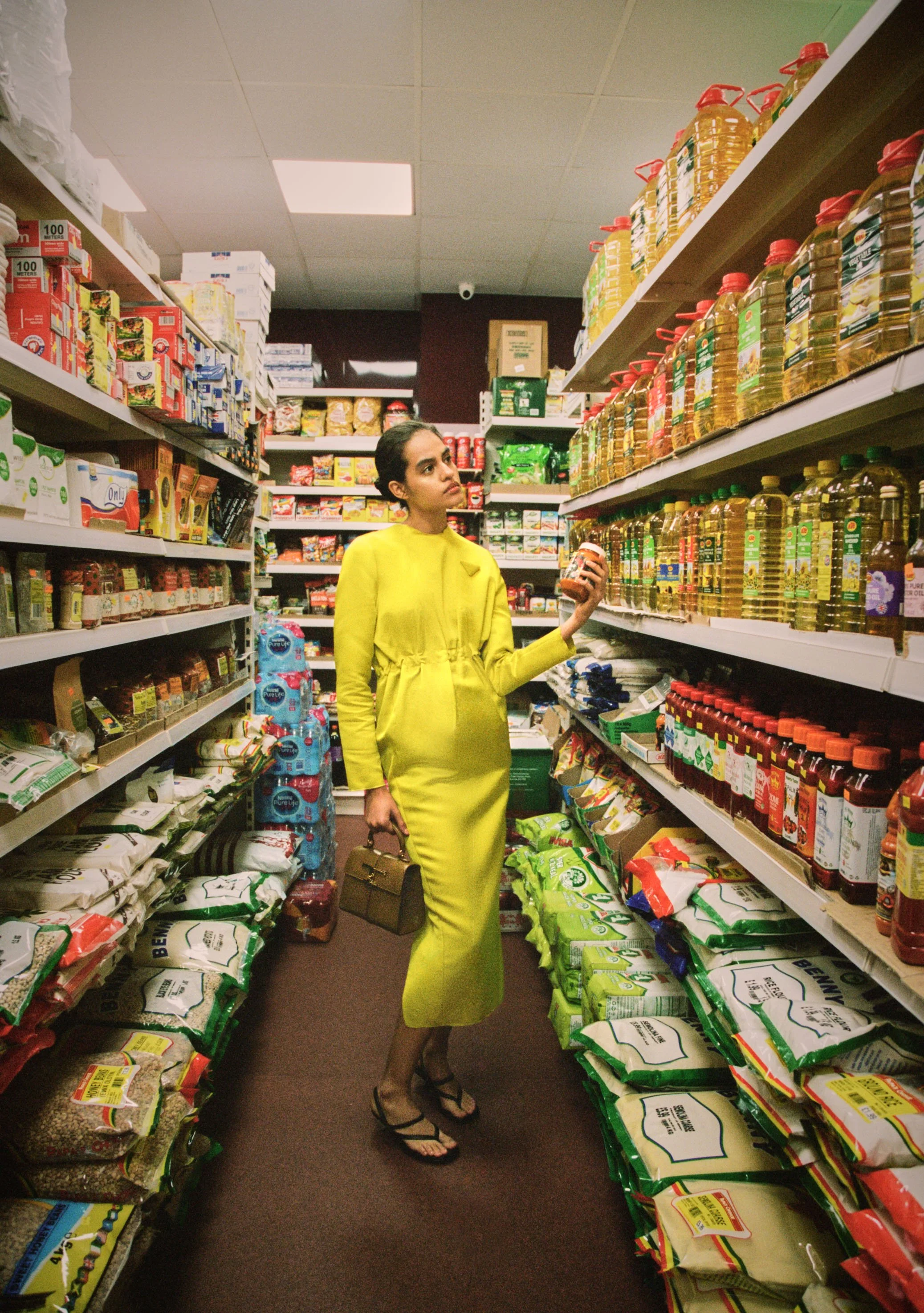 A woman in a bright yellow dress shopping in a grocery store aisle, holding a jar of food and a handbag, surrounded by shelves stocked with cooking oils, sauces, spices, and dried goods.