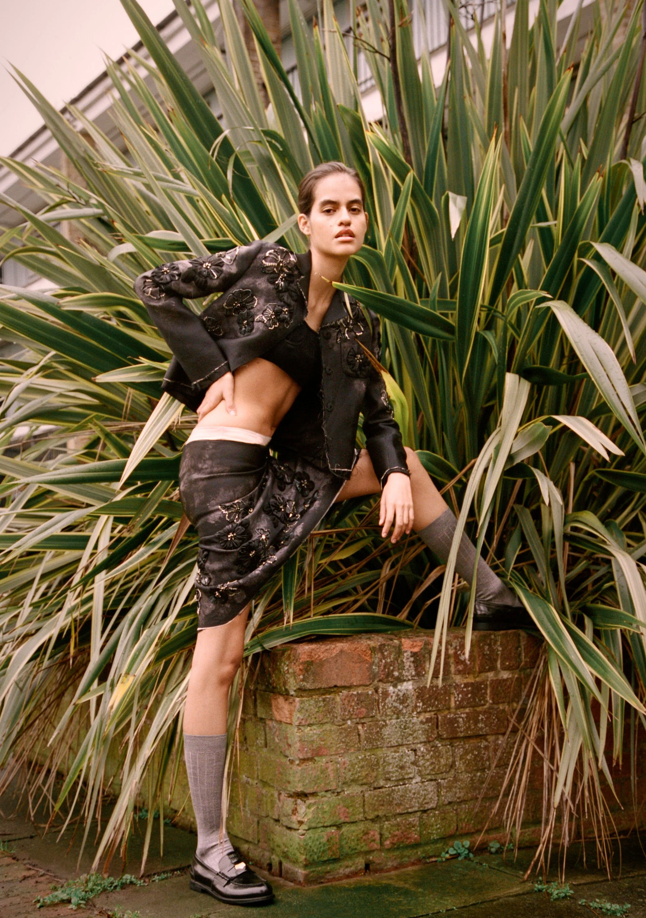 A young woman dressed in a black embroidered jacket and skirt, with one leg raised on a brick planter, standing amidst tall green plants inside a greenhouse.
