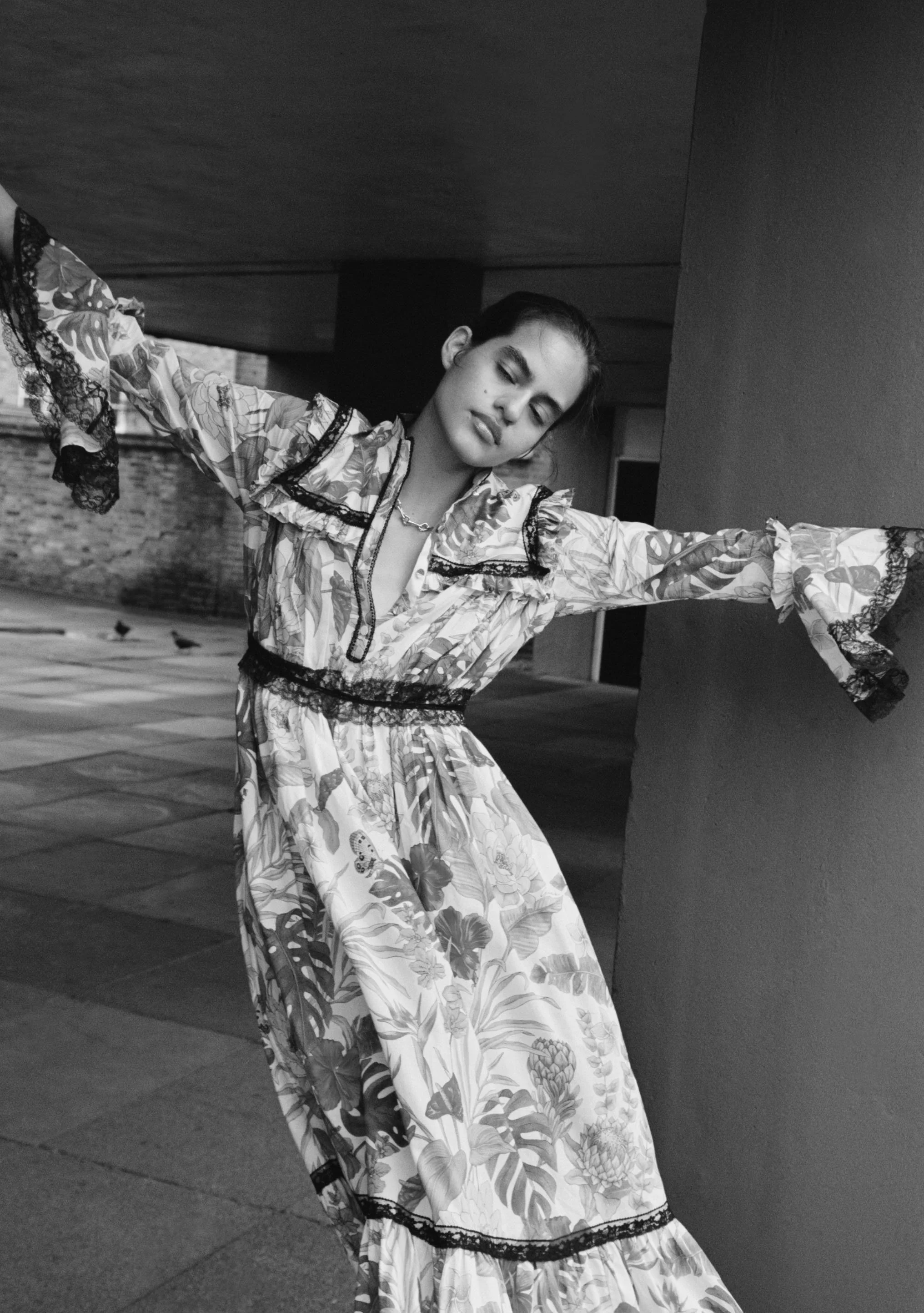 A woman in a floral dress with lace details, posing with arms outstretched in an urban setting, in black and white.
