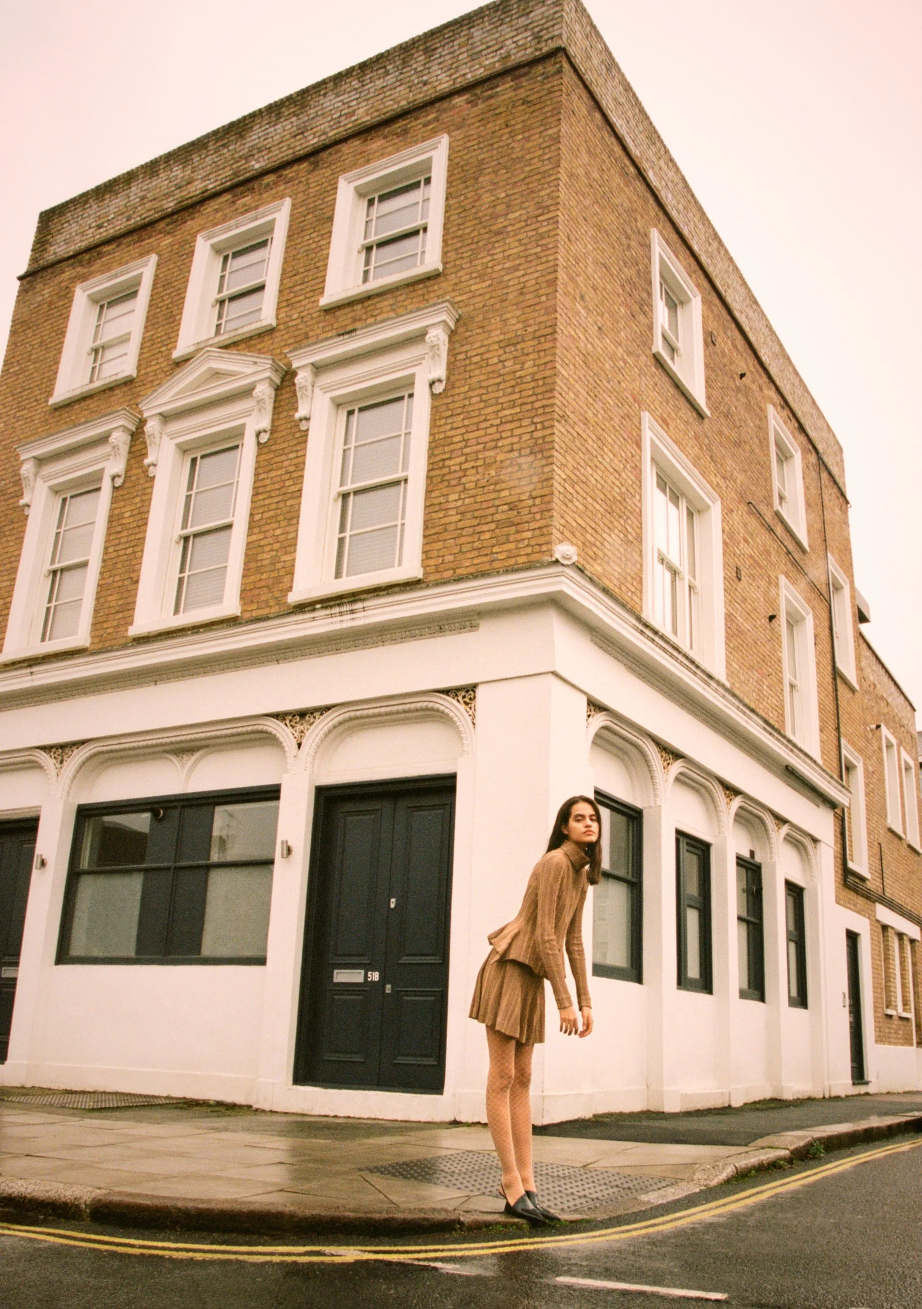 A young woman with dark hair wearing a tan dress and black heels stands on a city sidewalk in front of a corner building with a brick upper level and a white lower level featuring black doors and windows.