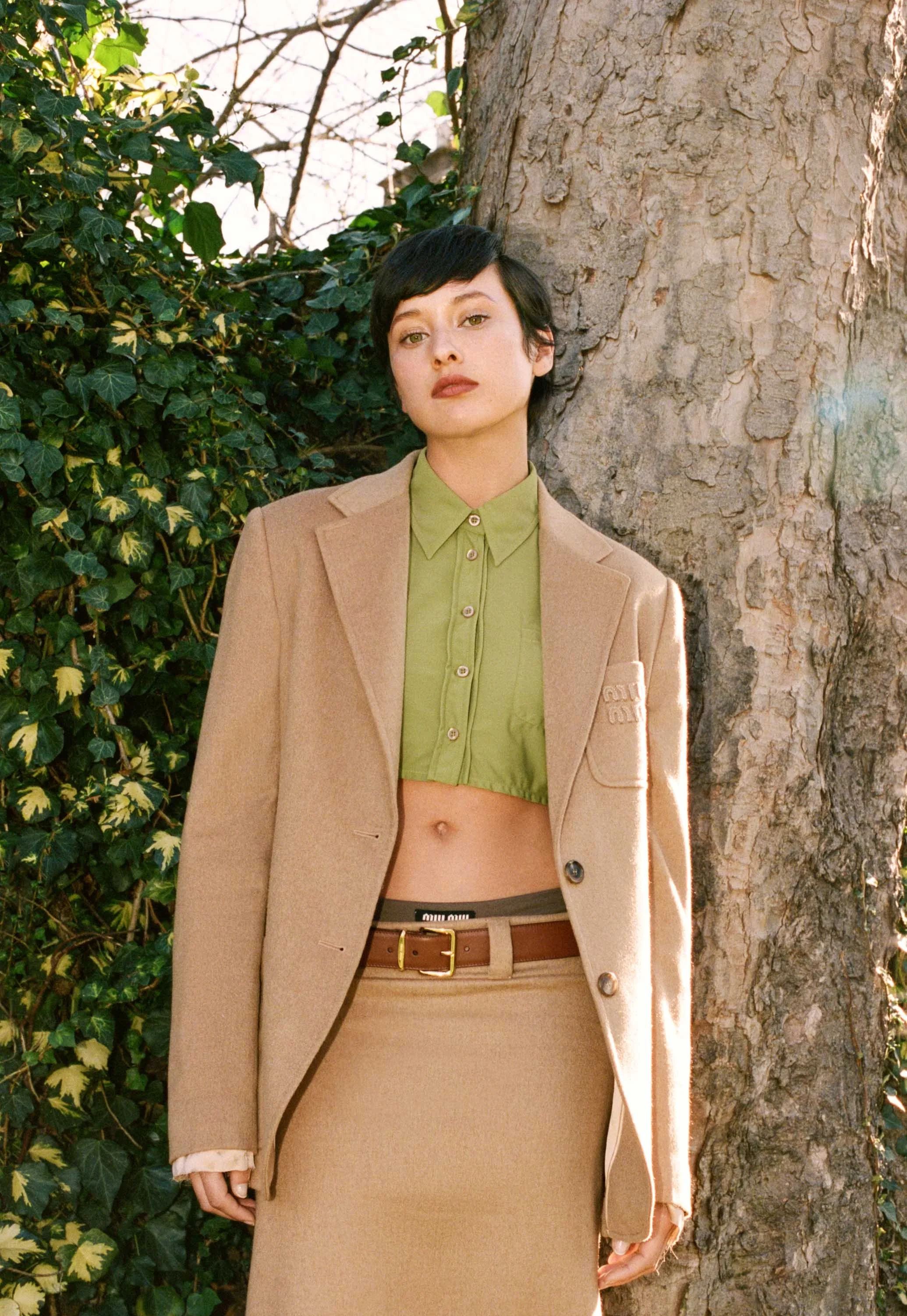 A woman with short dark hair and fair skin standing outdoors next to a large tree trunk, wearing a beige blazer, a green cropped shirt, and a matching beige skirt, with greenery in the background.