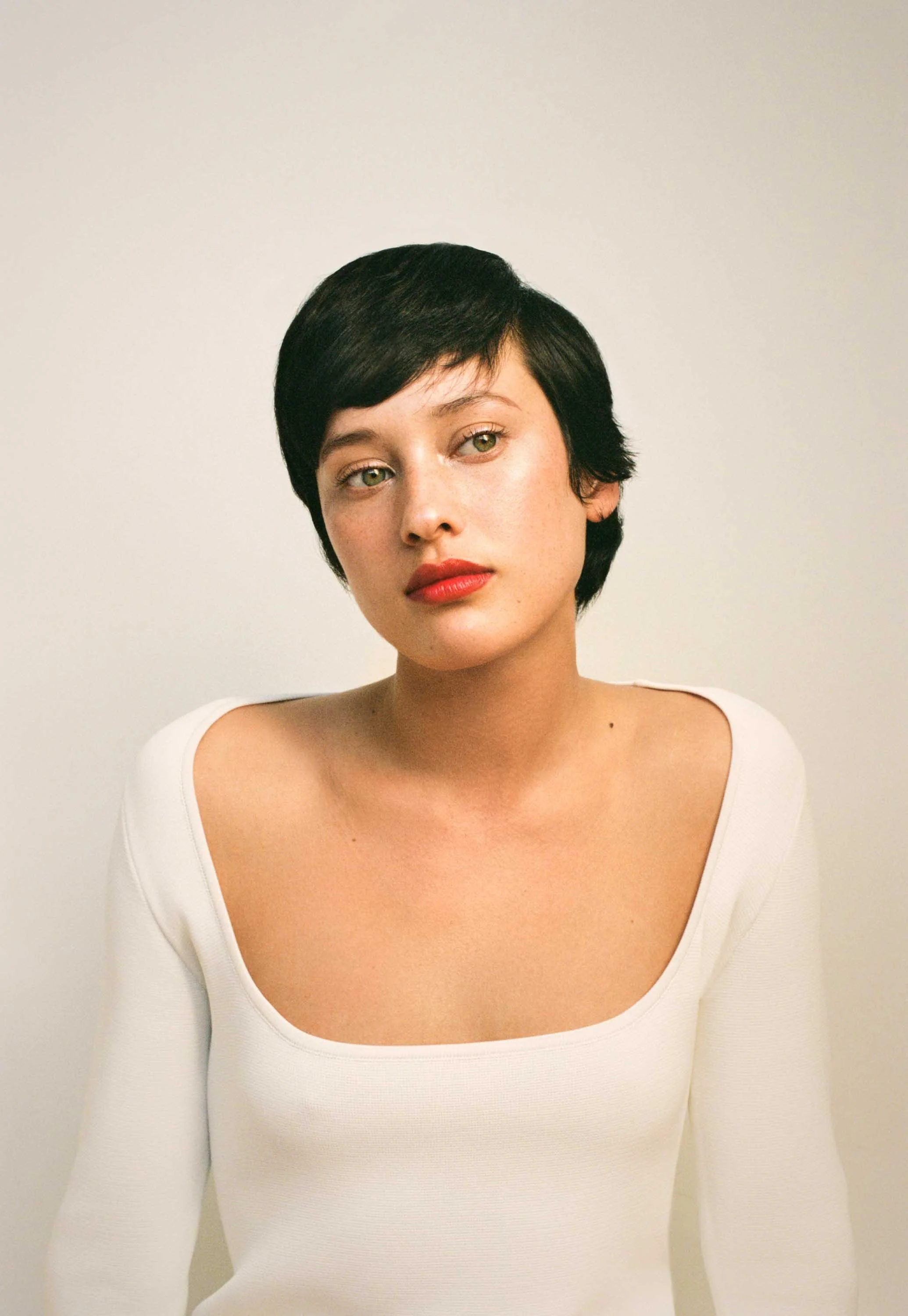A young woman with short black hair and red lipstick looks slightly to the side, wearing a white top with a rounded neckline against a plain, light-colored background.