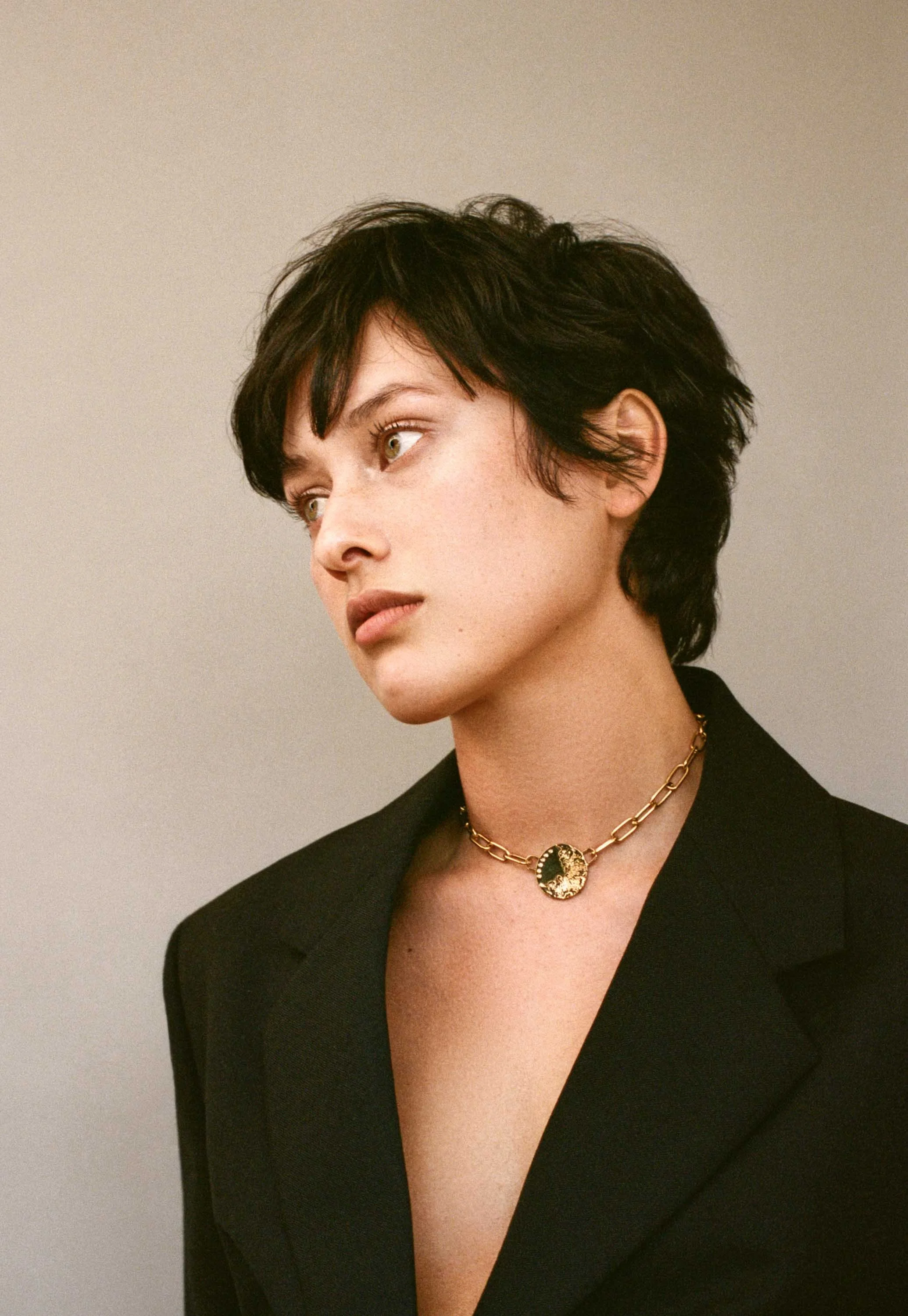 A woman with short dark hair, wearing a black blazer and a gold chain necklace with a decorative pendant, looking to the side against a plain background.