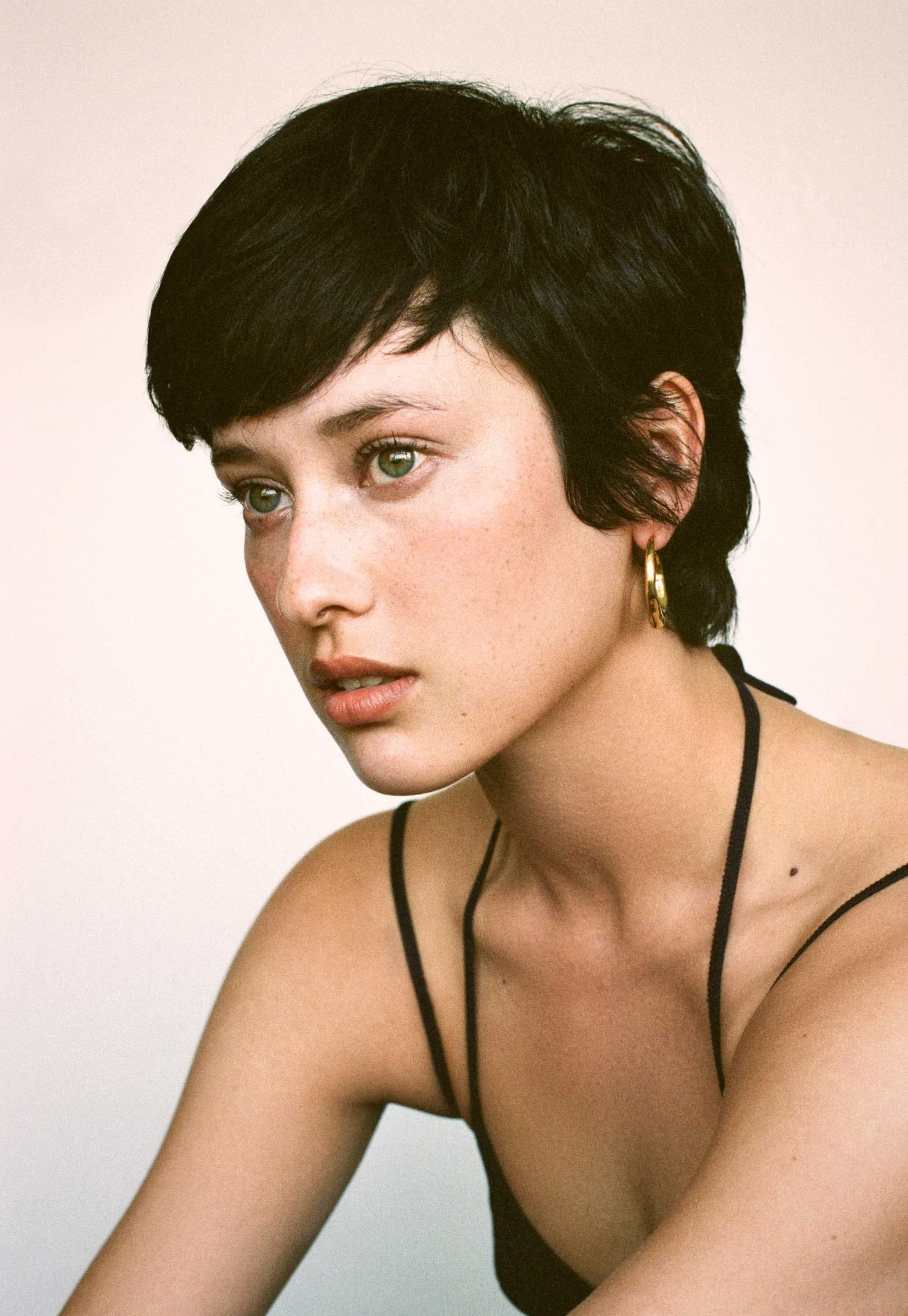 A woman with short black hair, green eyes, and light skin looking to the side. She is wearing gold hoop earrings and a black spaghetti strap top.