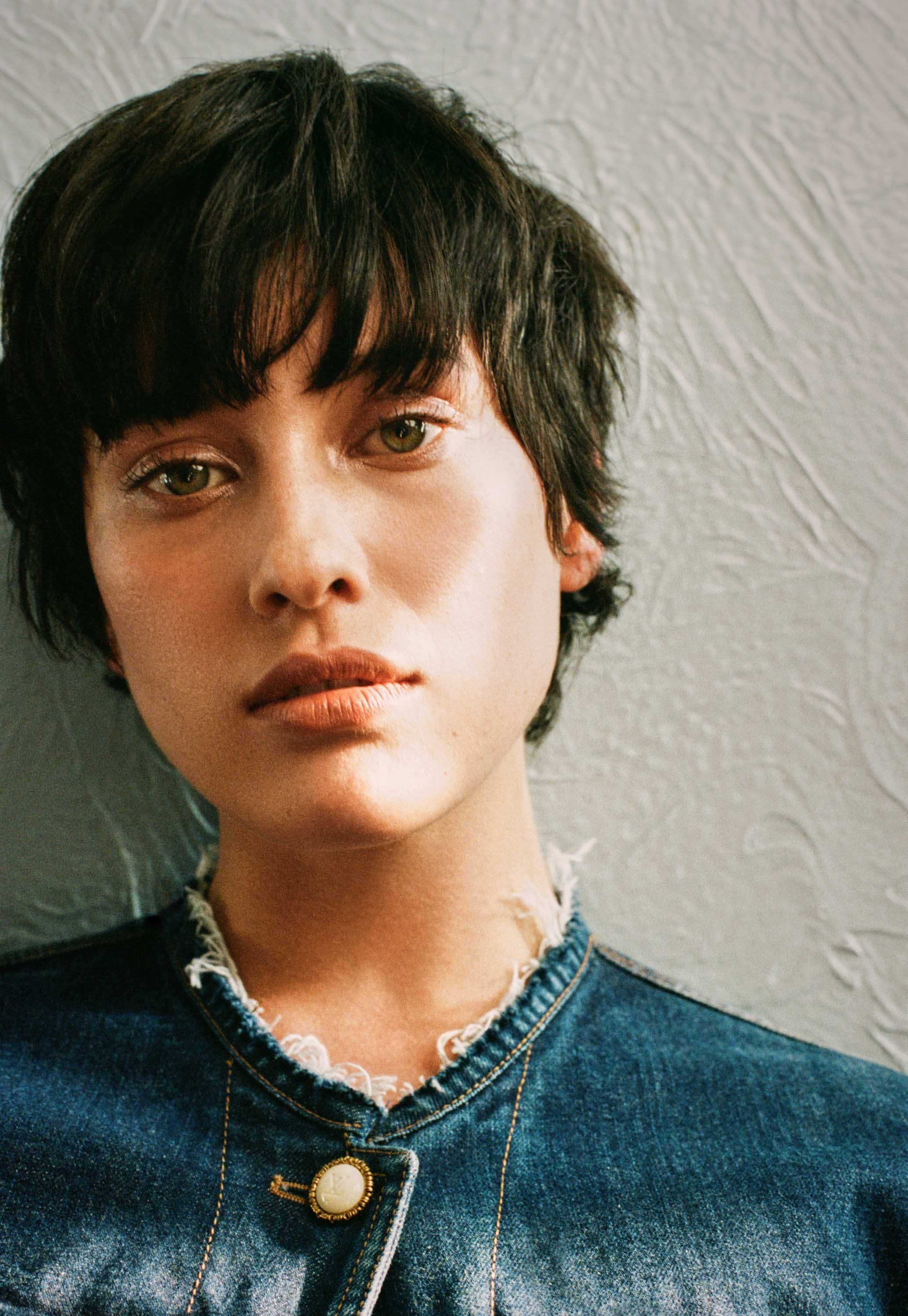 A woman with short, dark, tousled hair and green eyes, wearing a denim jacket with a frayed collar and a gold button, standing against a textured white wall.