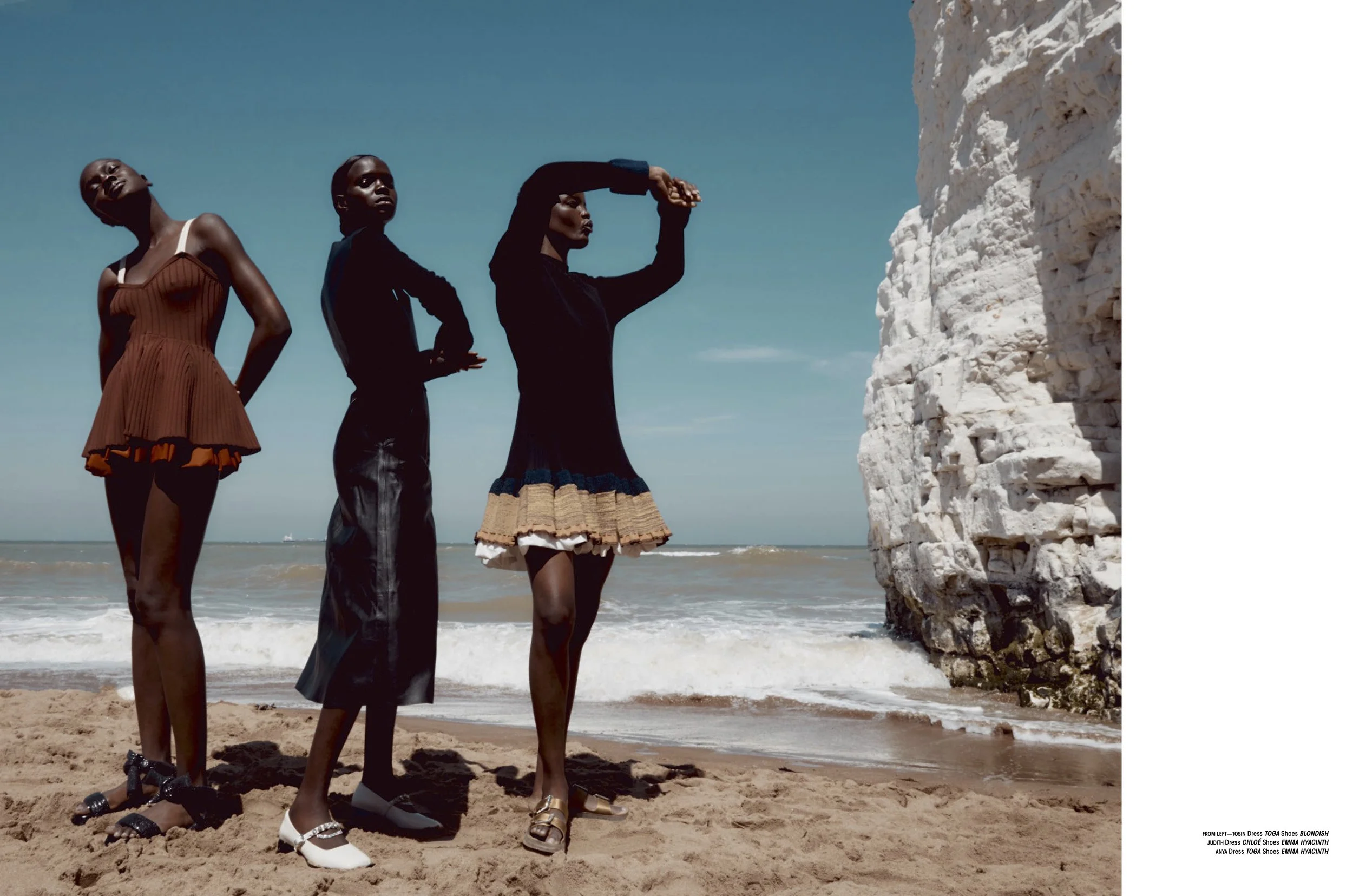 Three women posing on a sandy beach near a white rock cliff, wearing fashionable dresses and shoes, with the ocean and blue sky in the background.