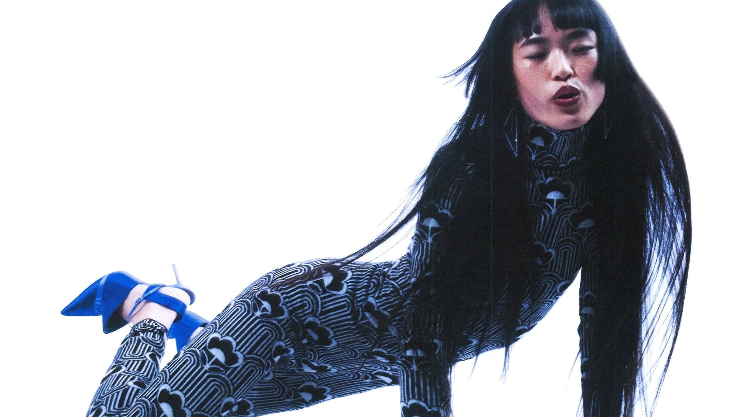 A woman with long black hair wearing a matching black and white patterned outfit and blue shoes, posing against a plain white background.