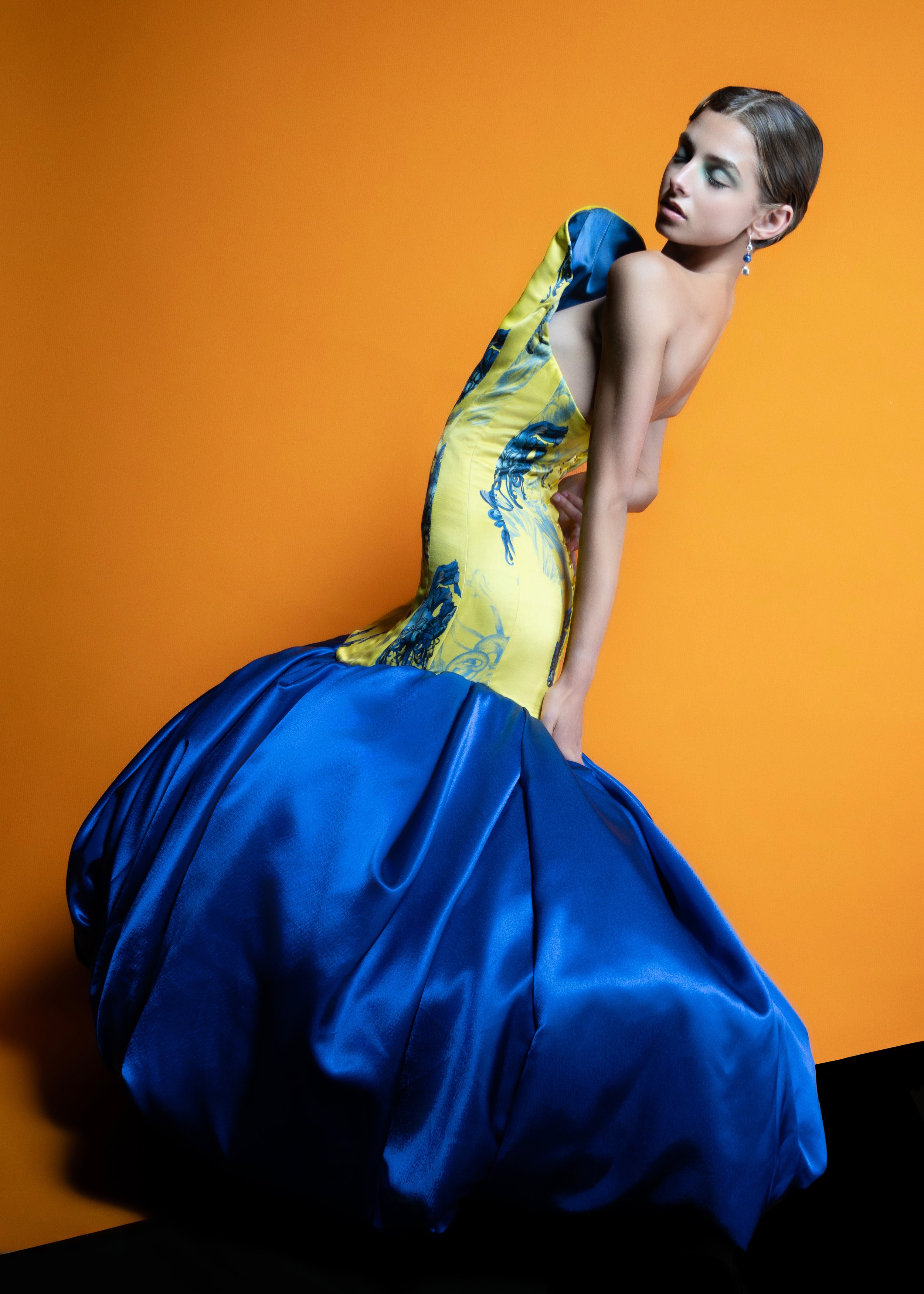 A woman in an elaborate yellow and blue gown poses against an orange background.