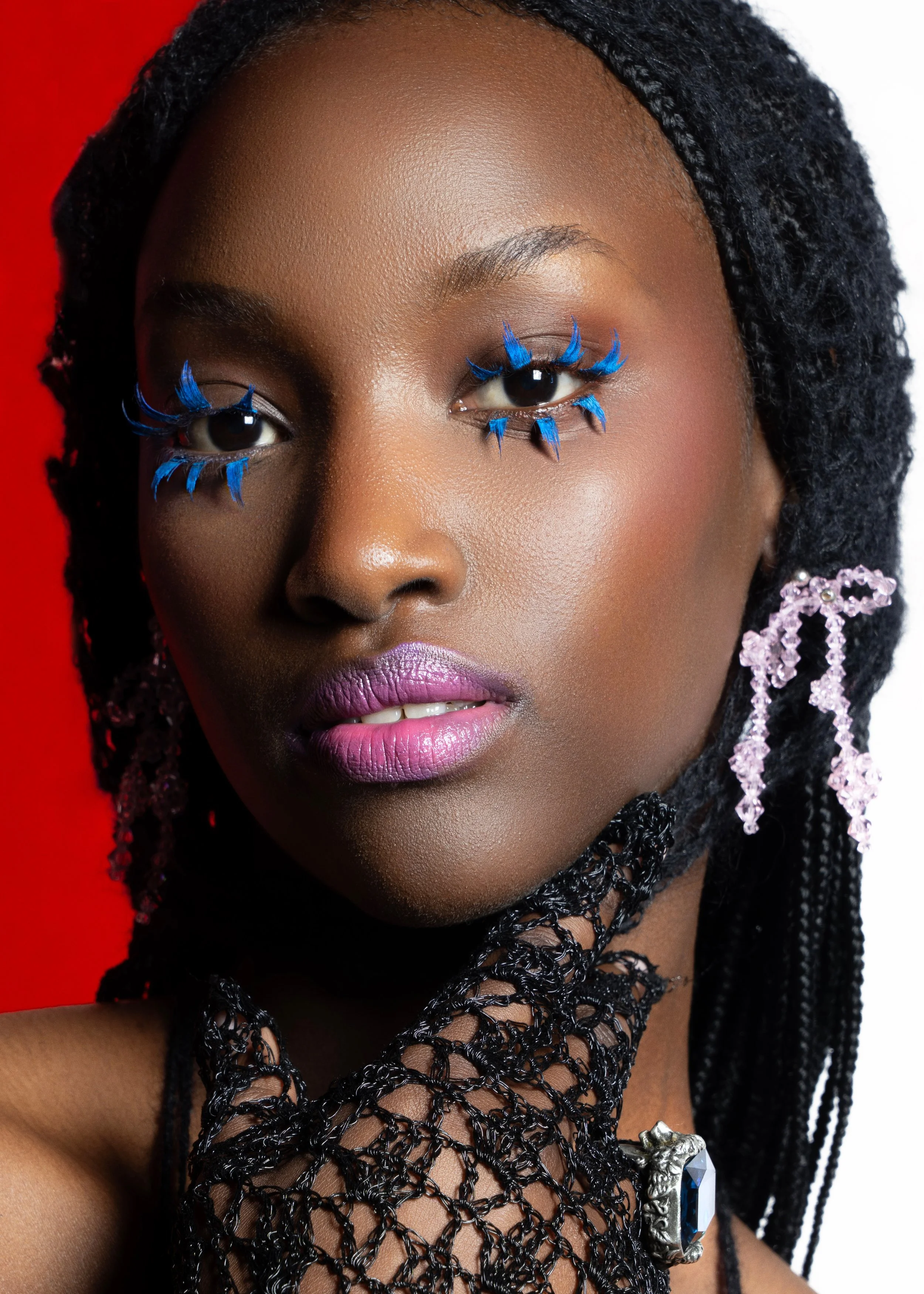 Close-up of a woman with dark skin, wearing blue feathered eyelashes, pink lipstick, large earrings, and a black woven necklace with a large gemstone ring on her finger, against a red and white background.