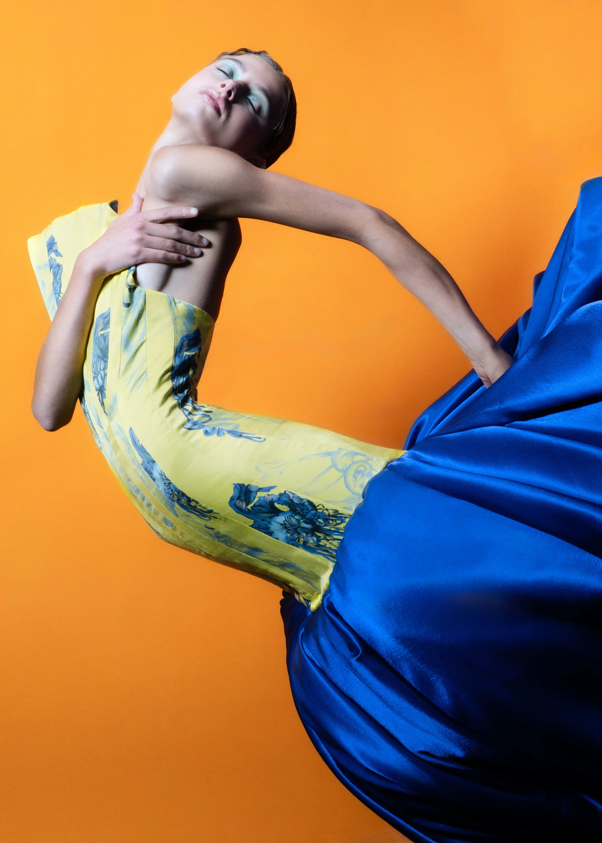 A woman with closed eyes and pastel makeup, wearing a yellow and blue patterned dress and a full-length blue satin skirt against an orange background.