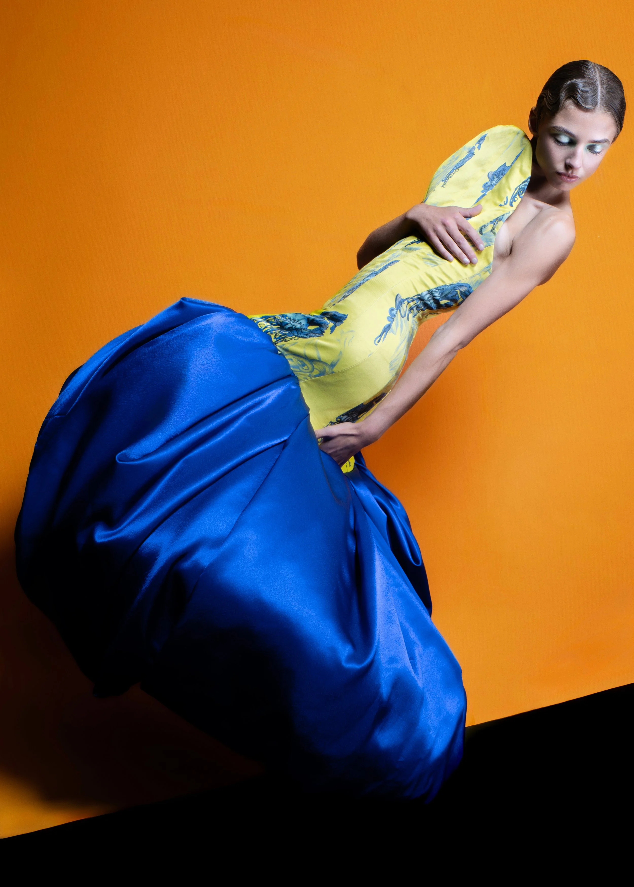 A woman wearing a yellow top with blue floral designs and a voluminous blue satin skirt, holding a matching yellow shawl with blue patterns, standing against an orange background.