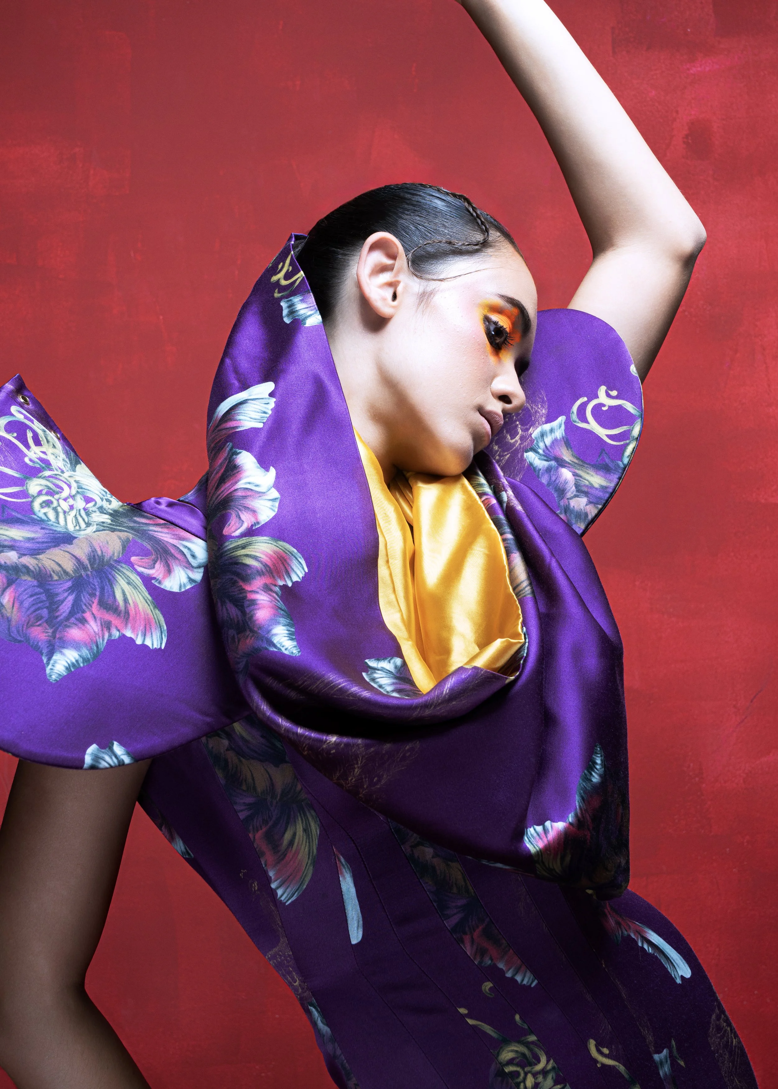 A woman posing with her eyes closed, wearing a purple floral kimono-like garment with yellow lining and vibrant makeup, against a red textured background.