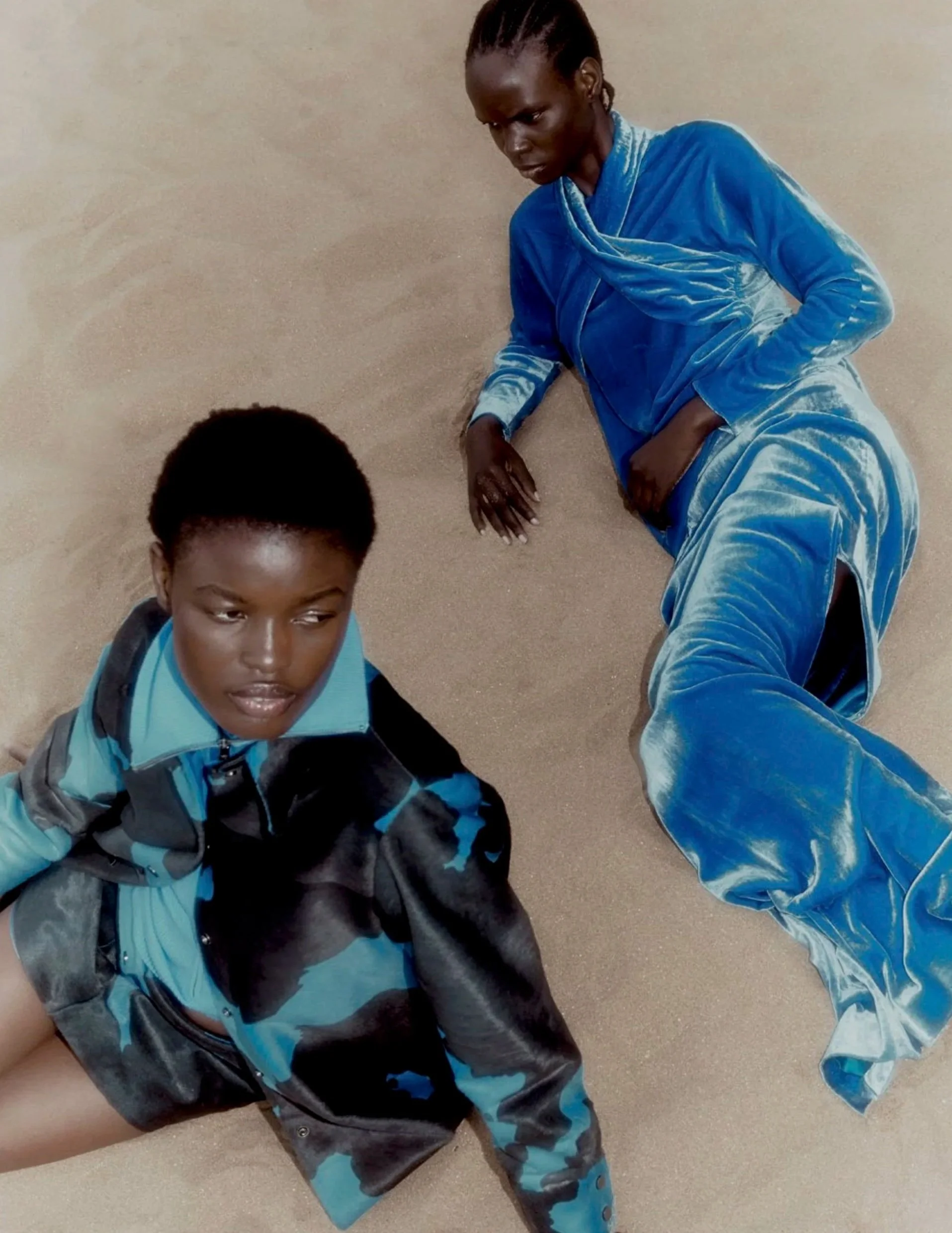 Two models, one in a camouflage jacket and shorts, and the other in a blue velour tracksuit, lying on sandy ground.