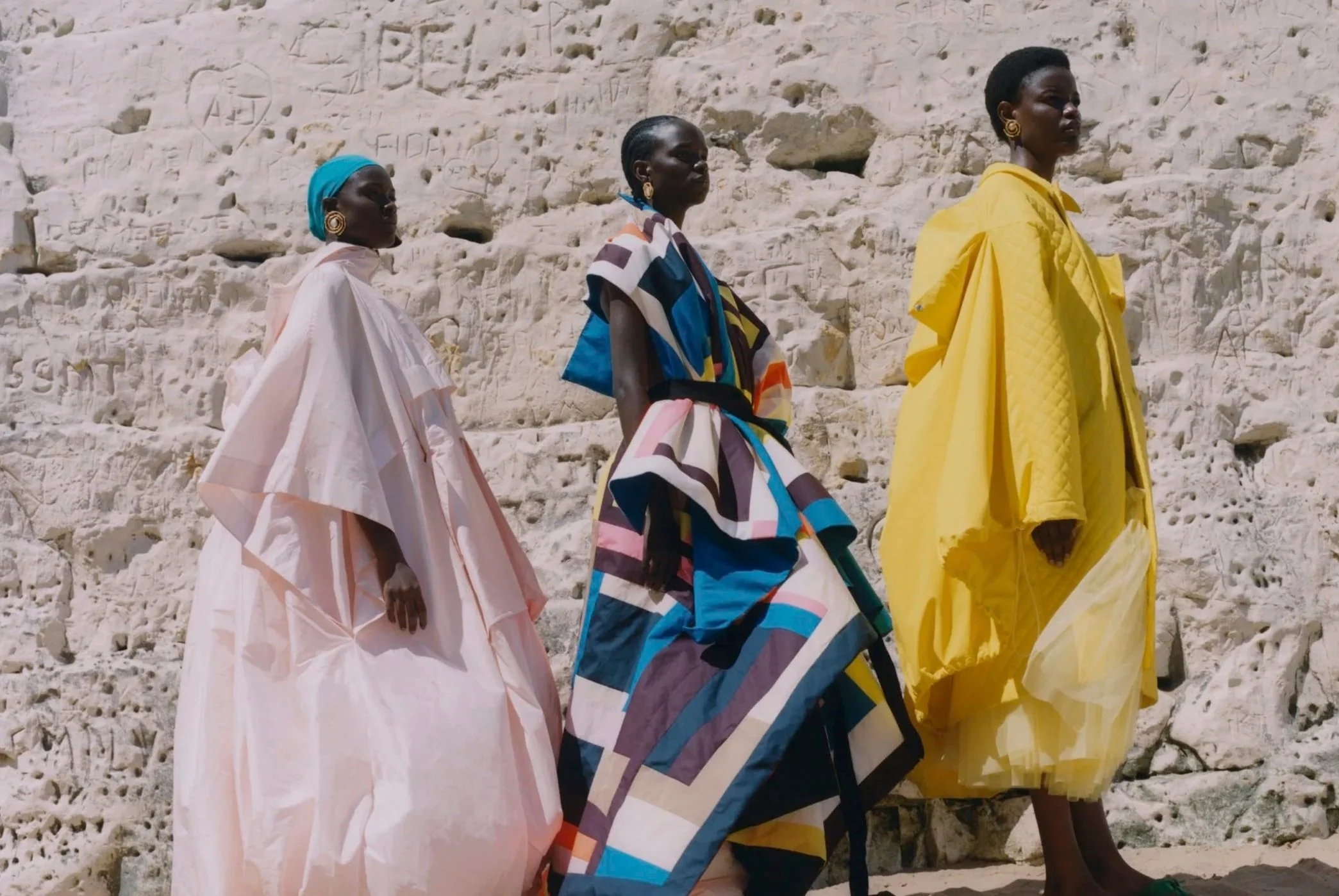 Three women standing outdoors against a stone wall, wearing colorful, flowing garments in pink, multicolored geometric pattern, and yellow, with the text "On the Sea" by the New York Times appearing vertically on the right side of the image.