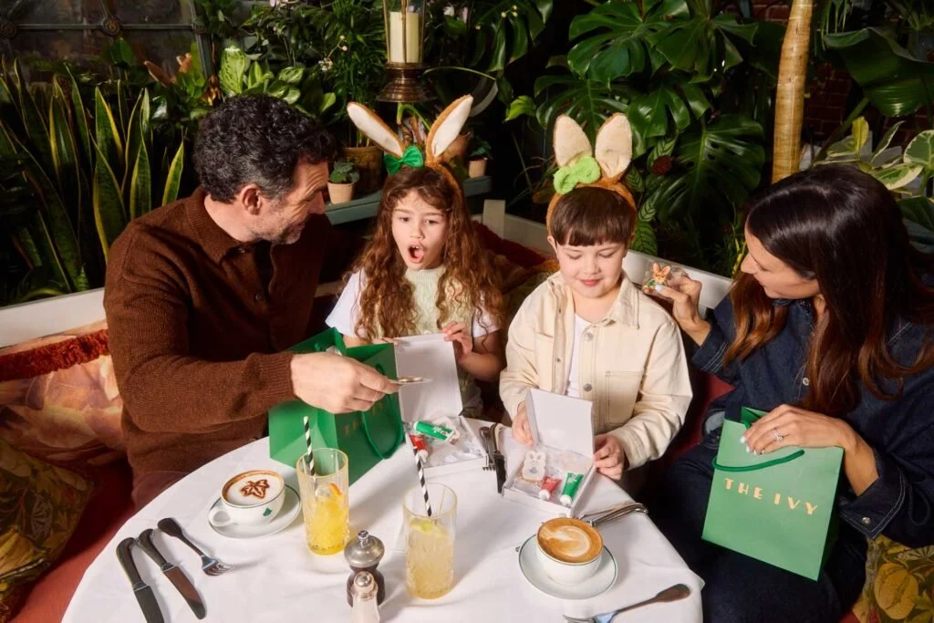 Family celebrating Easter at a restaurant, with children wearing bunny ear headbands, opening gifts, and sitting at a table with drinks and desserts.