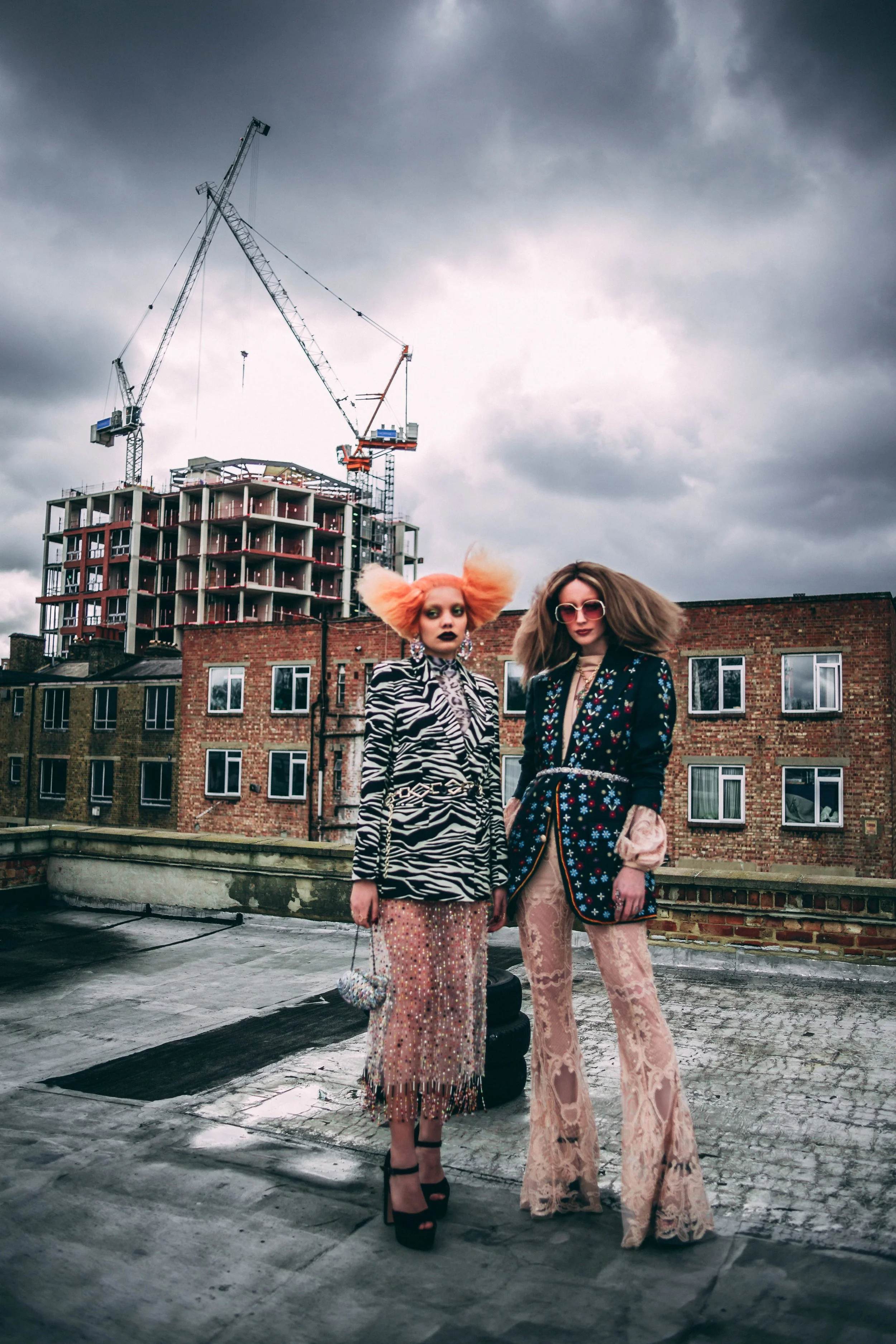 Two women with dramatic fashion styles standing on a rooftop with a construction site and cloudy sky behind them.