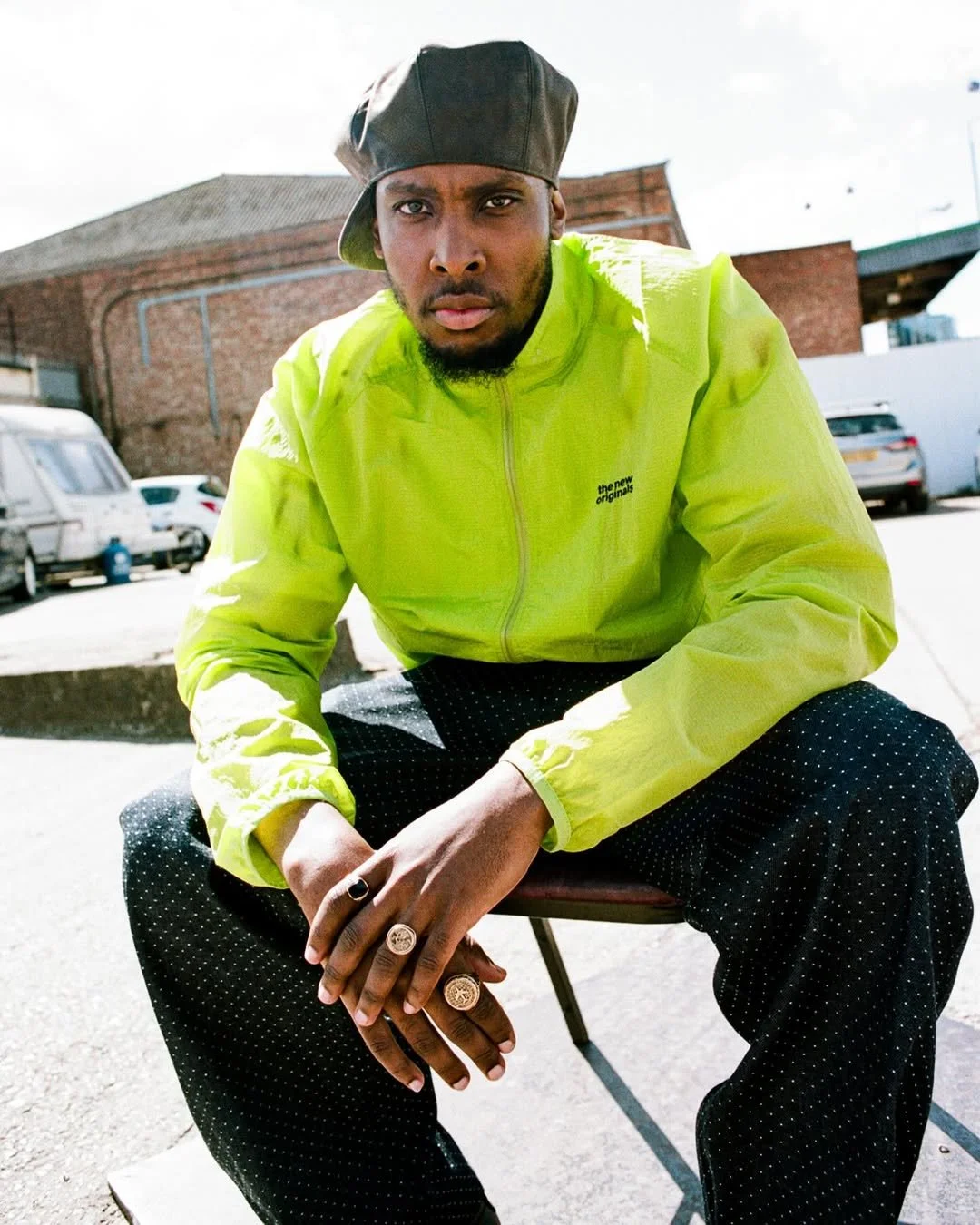 A man wearing a black cap backwards and a bright yellow jacket with text on the chest, sitting on a chair outdoors with cars and brick buildings in the background.