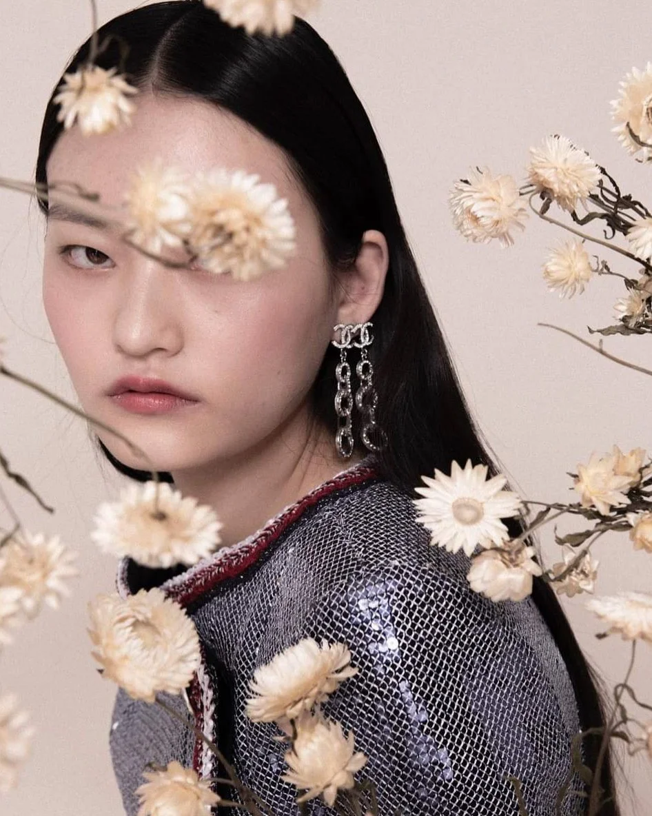 A woman with long black hair wearing a sequin dress and statement earrings, surrounded by cream-colored flowers.