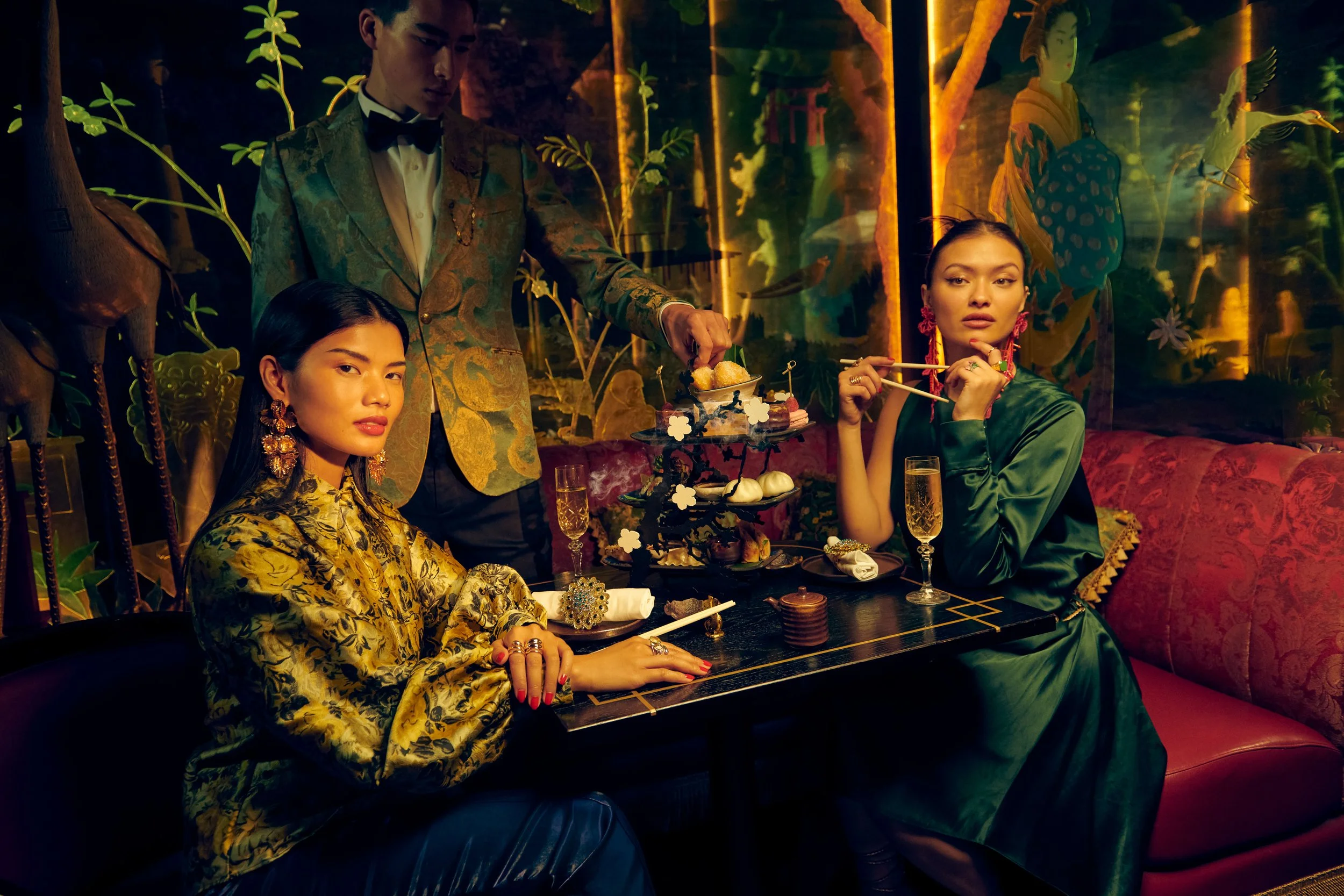 Inside a dimly lit, luxurious restaurant, two women with elaborate jewelry and elegant dresses sit at a table while a man in a patterned suit serves them a three-tiered tray of desserts. The background features vibrant, artistic wall decorations with animal and nature motifs.
