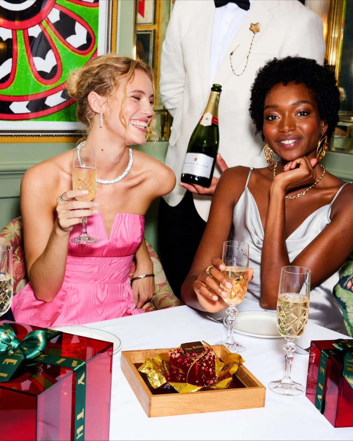 Two women celebrating at a party, dressed in elegant attire, holding glasses of champagne, with gift boxes and a decorated cake on the table.