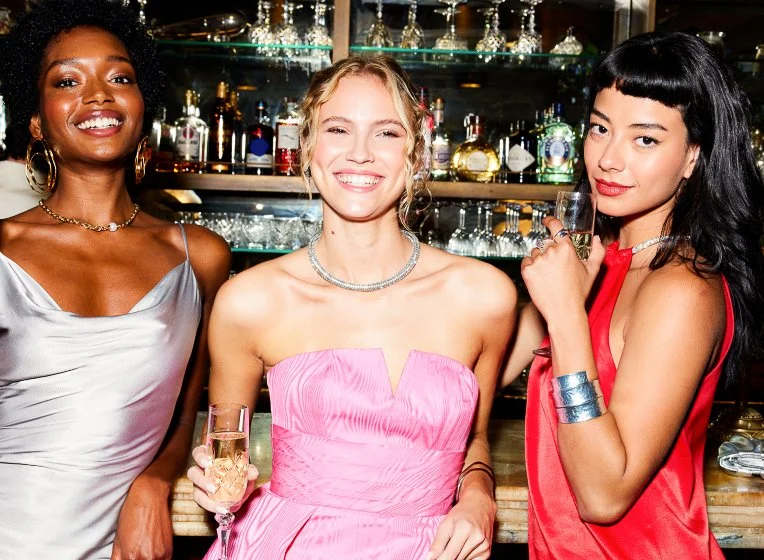 Three women at a bar holding glasses of champagne, smiling and dressed in stylish evening wear.