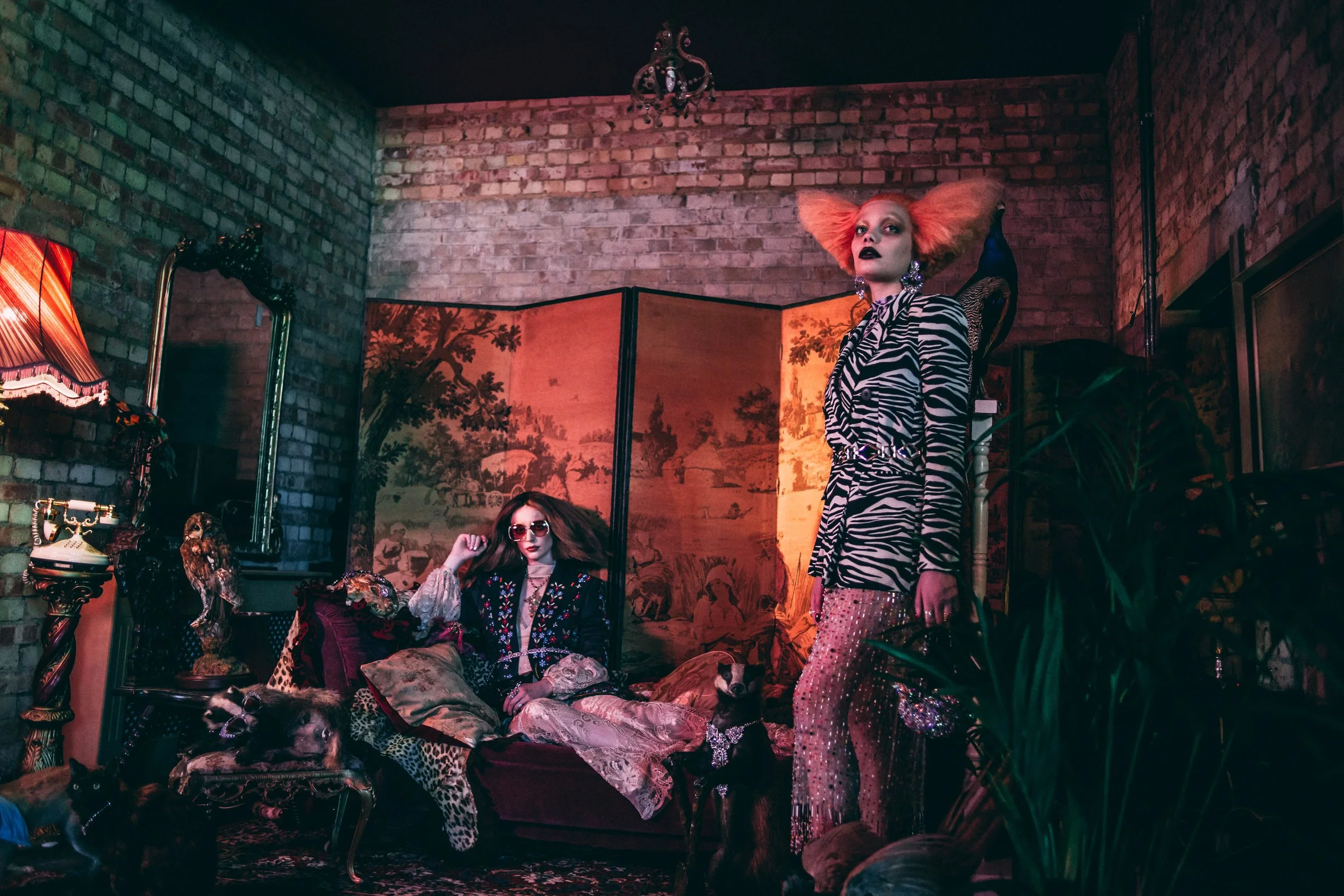 Two women with eccentric fashion and bright hair in a vintage styled room with exposed brick walls, ornate lamps, and a decorative screen, surrounded by dogs and vintage decor.