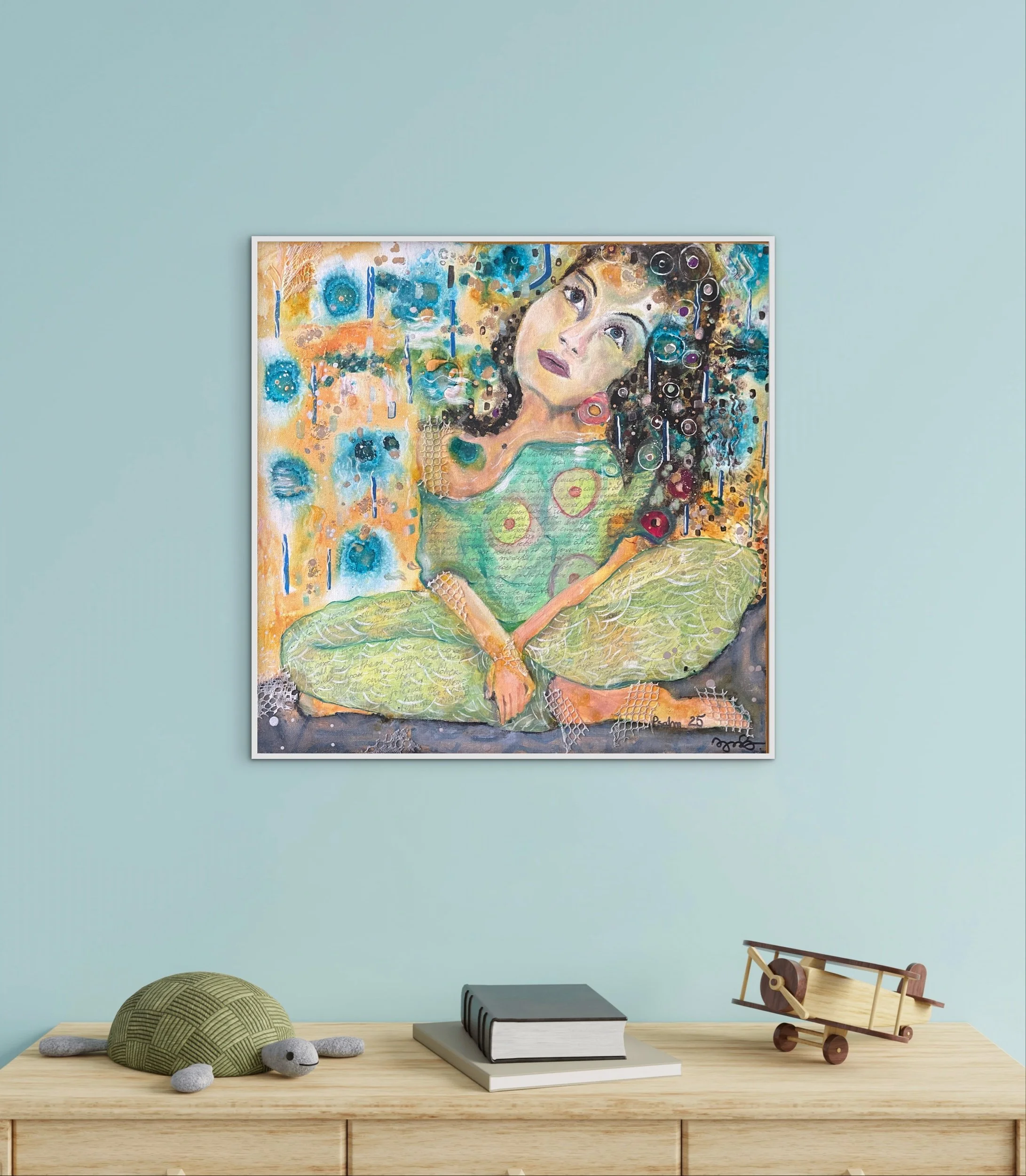 A wooden desk with a gray book, a large black and white hardcover , a plush turtle toy, a plush gray mouse toy, a small wooden toy airplane, and a light green decorative ball on it. Framed abstract artwork hangs on a pale blue wall above, and a black box or electronic device is on the desk.