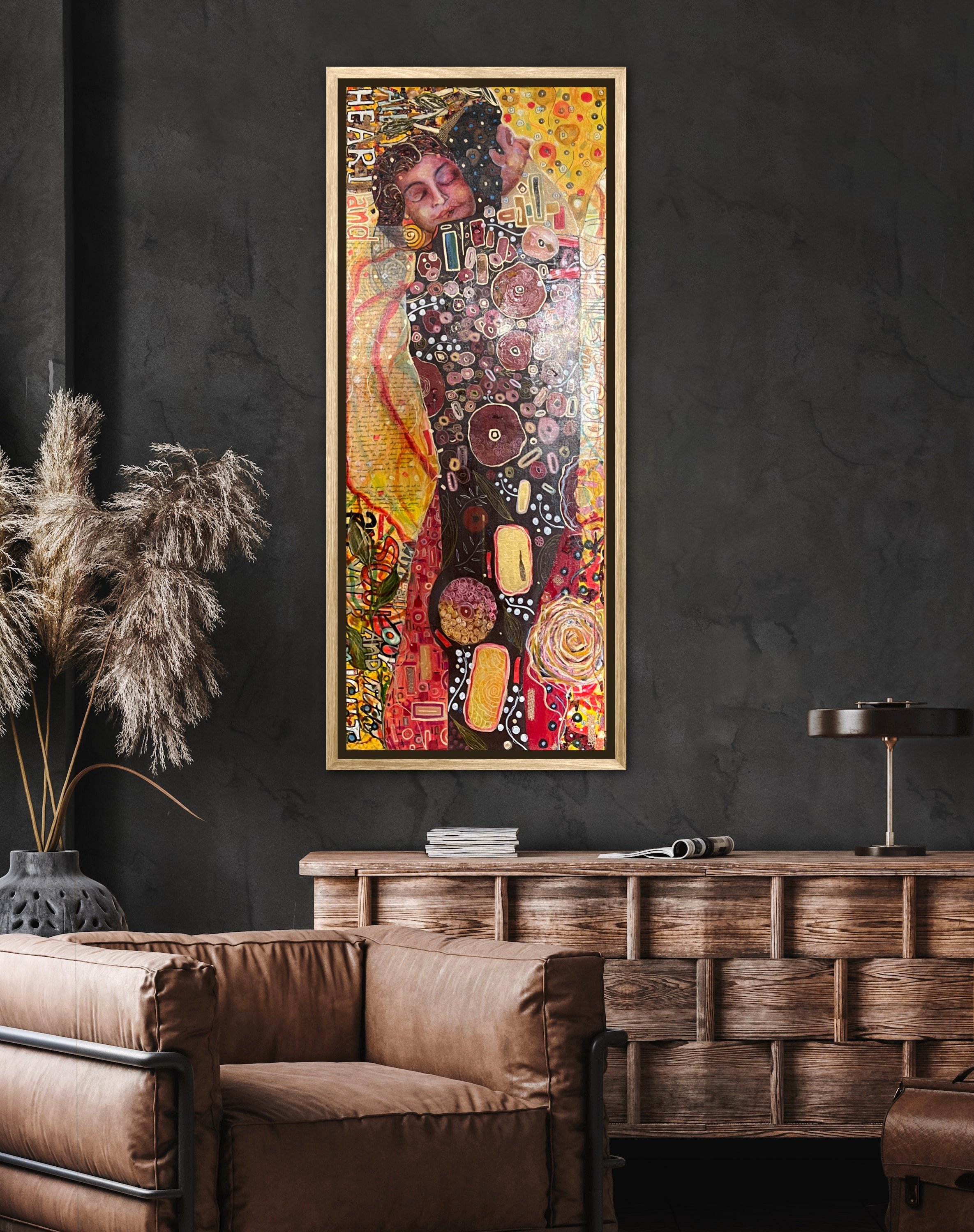 Interior of a modern living room with a brown leather sofa, a wooden sideboard, a black table lamp, a decorative vase with dried pampas grass, and a large colorful abstract painting on a dark wall.