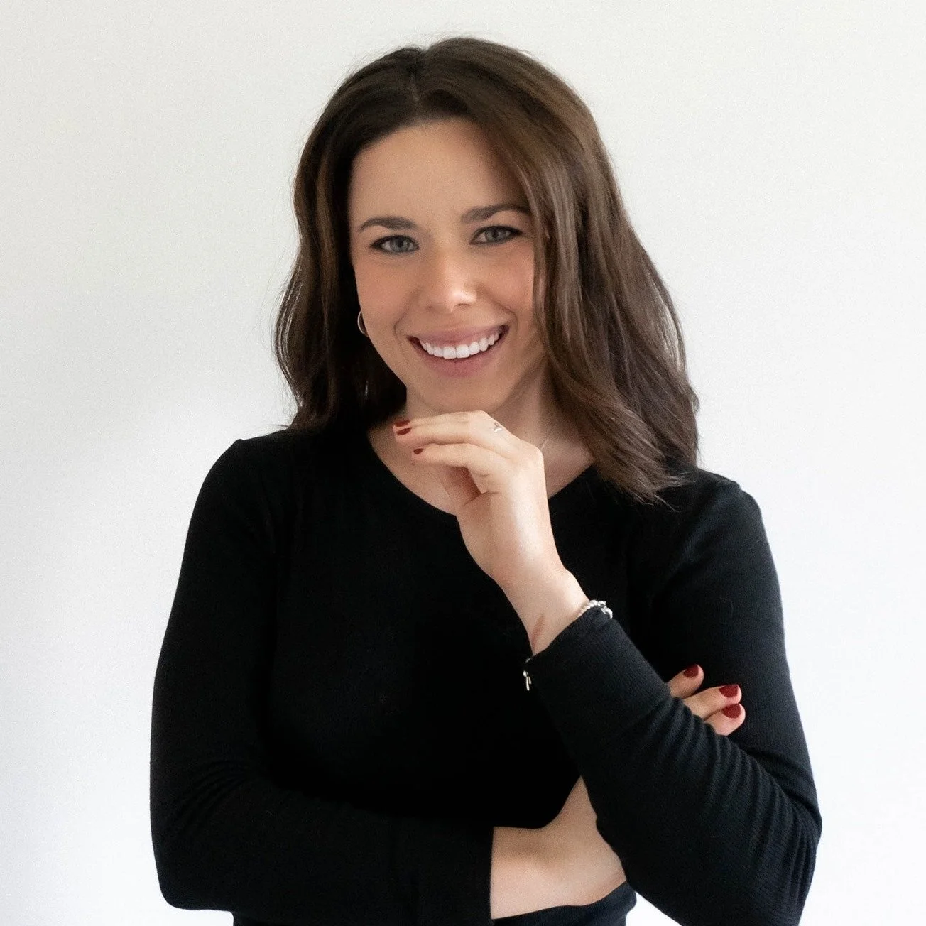 A woman with shoulder-length brown hair, wearing a black long-sleeve top, smiling with her hand near her chin, standing against a plain white background.