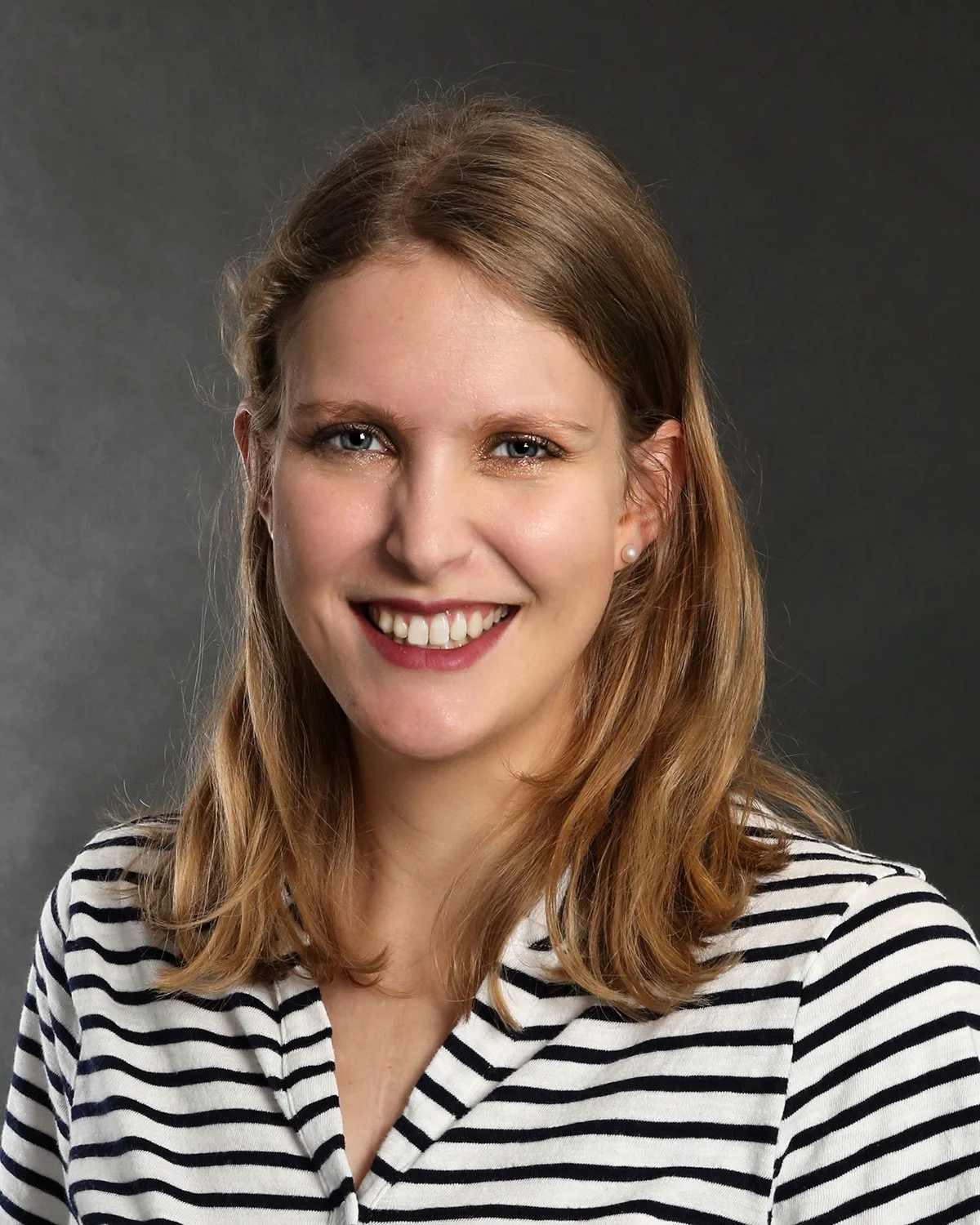 A woman with shoulder-length light brown hair, blue eyes, and fair skin, smiling and wearing a black and white striped shirt against a dark gray background.