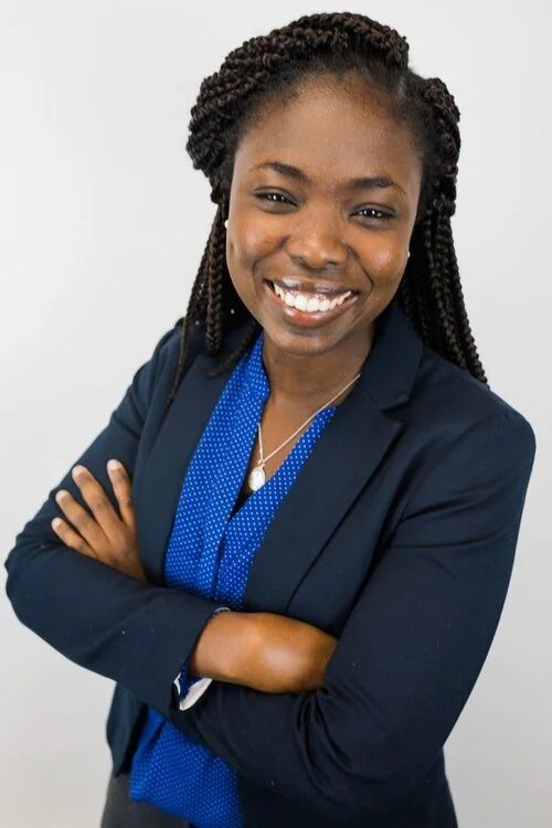 Professional woman with braided hair smiling confidently, wearing a navy blazer and blue blouse, arms crossed against a plain background.