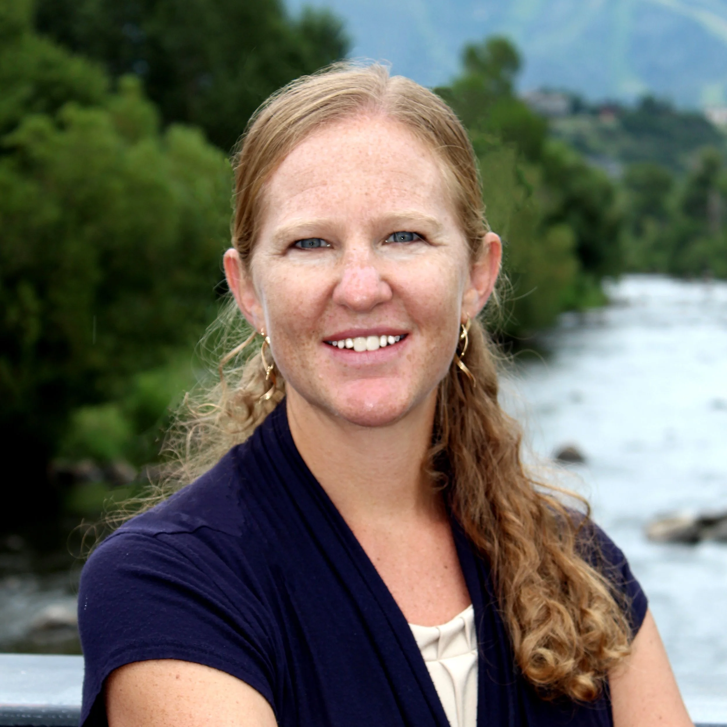 Portrait of a woman with red hair and freckles smiling outdoors with a river and green trees in the background.