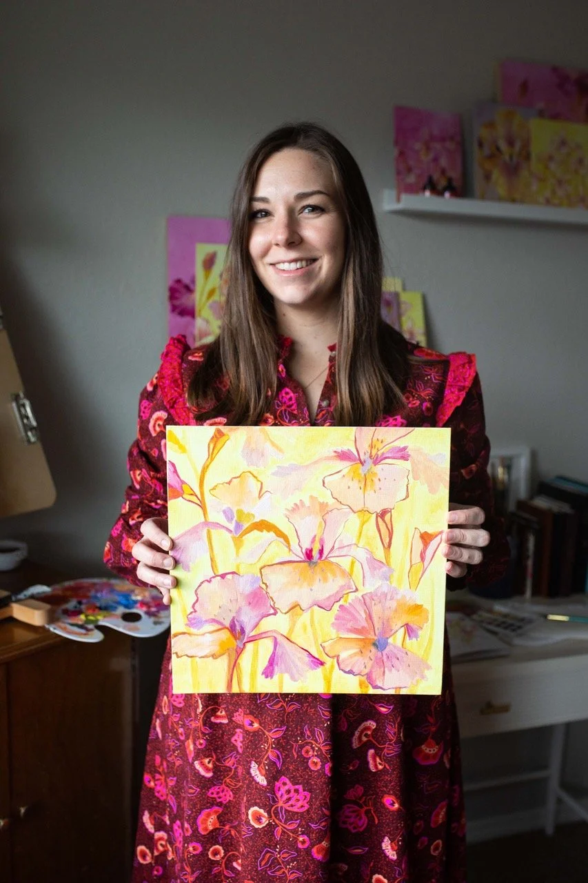 A woman with long brown hair smiling and holding a watercolor painting of pink and orange flowers. She is in an indoor art studio with paintings and art supplies in the background.