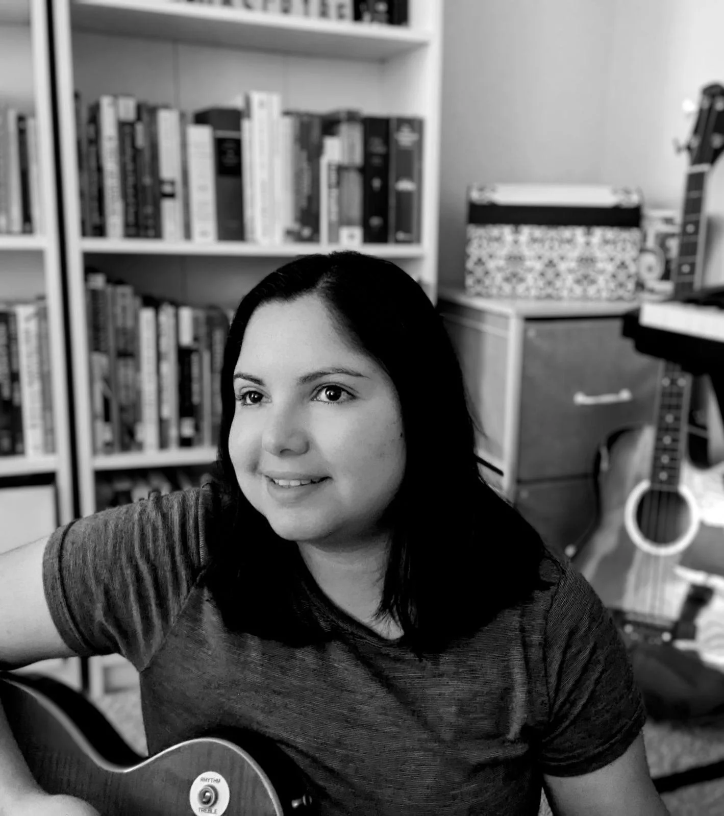 A woman with black hair smiling while playing a guitar in a room with bookshelves, a desk, a box, and a guitar.