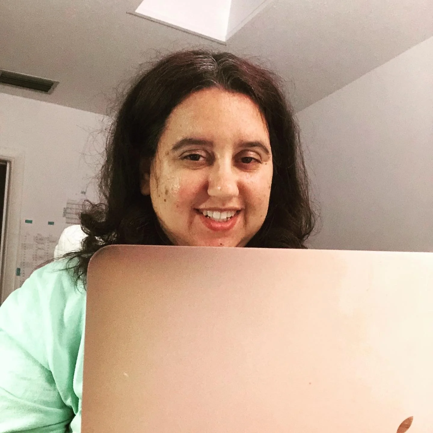 A woman with dark brown, wavy hair smiling while looking at a laptop in a well-lit room.