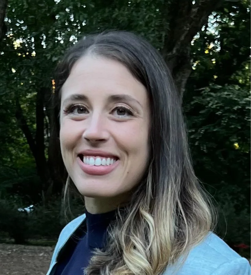 A young woman with long, wavy brown and blonde hair smiling outdoors in front of trees.
