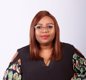Portrait of a woman with straight, shoulder-length hair wearing glasses, a black vest, and a colorful patterned shirt against a plain light background.
