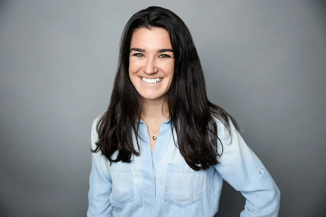 A woman with long dark hair smiling, wearing a light blue button-up shirt, standing against a gray background.