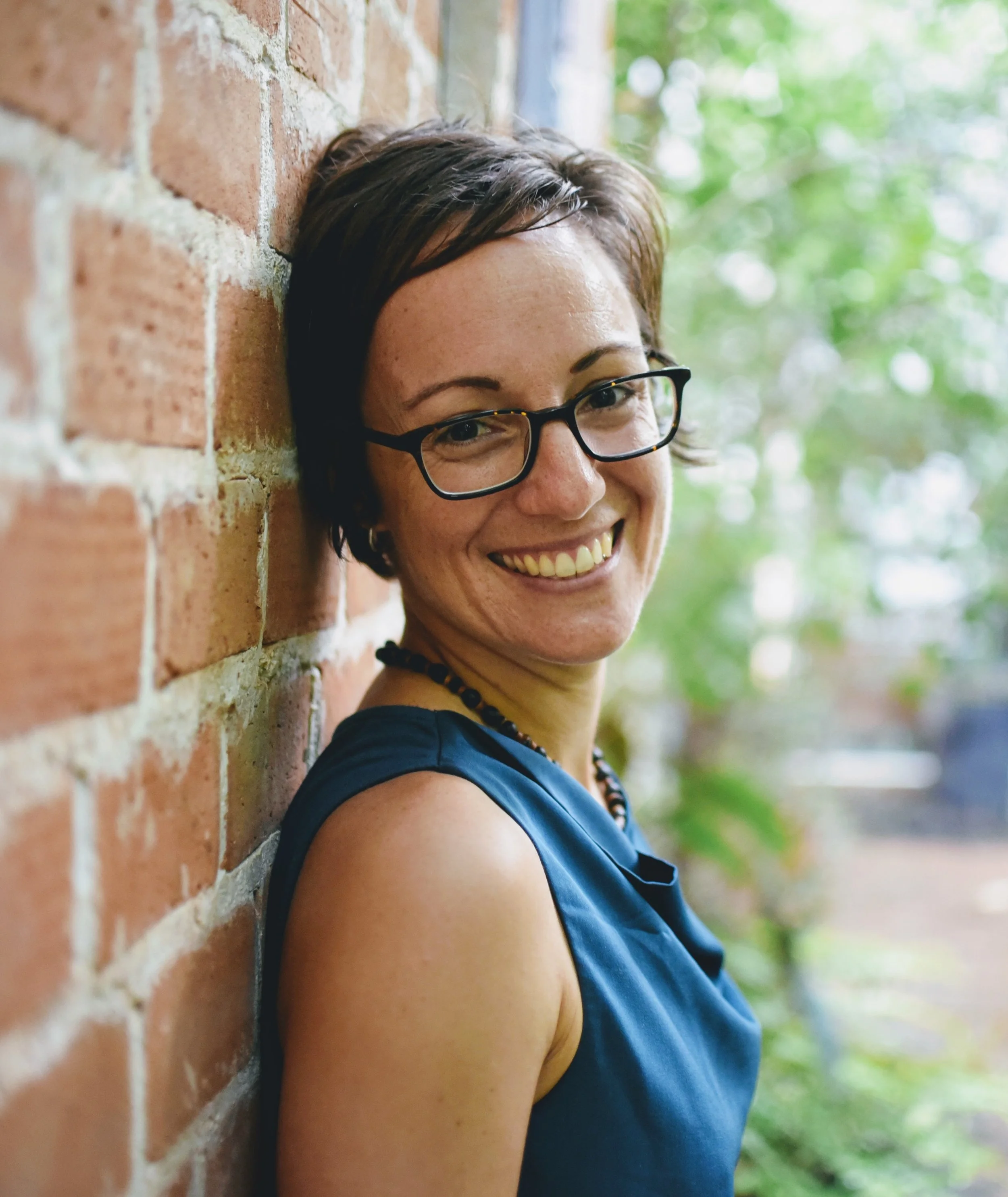 A woman with short dark hair and glasses leaning against a brick wall, smiling outdoors with greenery in the background.