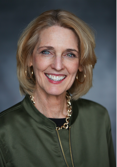 Headshot of a smiling middle-aged woman with shoulder-length blonde hair, wearing gold hoop earrings, a gold chain necklace, and a green jacket with a black top underneath.