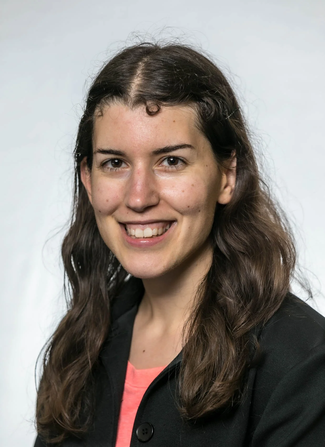 A woman with long brown wavy hair, smiling, wearing a black jacket and a pink shirt against a plain light background.