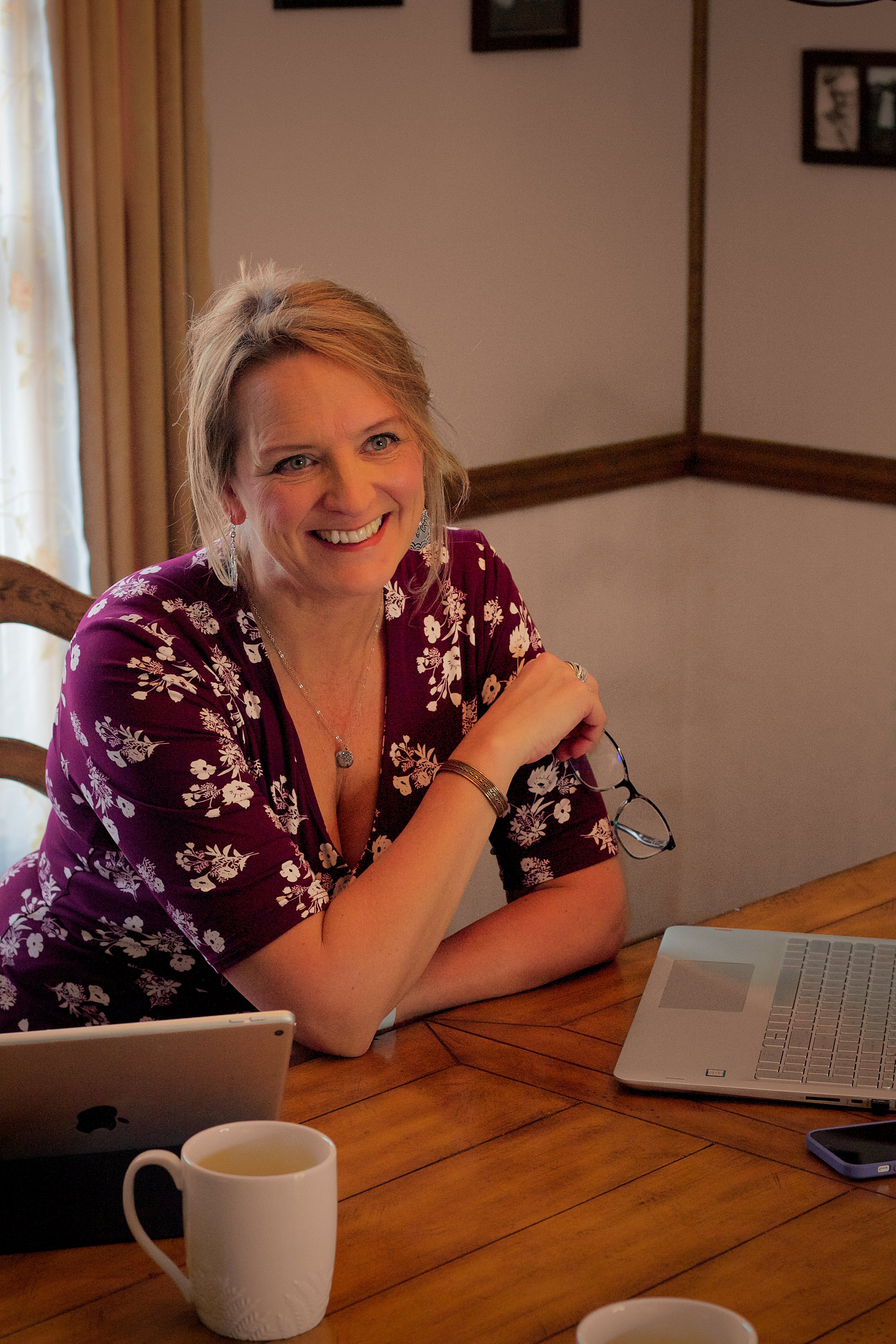 A smiling woman with blonde hair wearing a purple floral blouse, sitting at a wooden table with a laptop, a tablet, a smartphone, and two cups of tea.