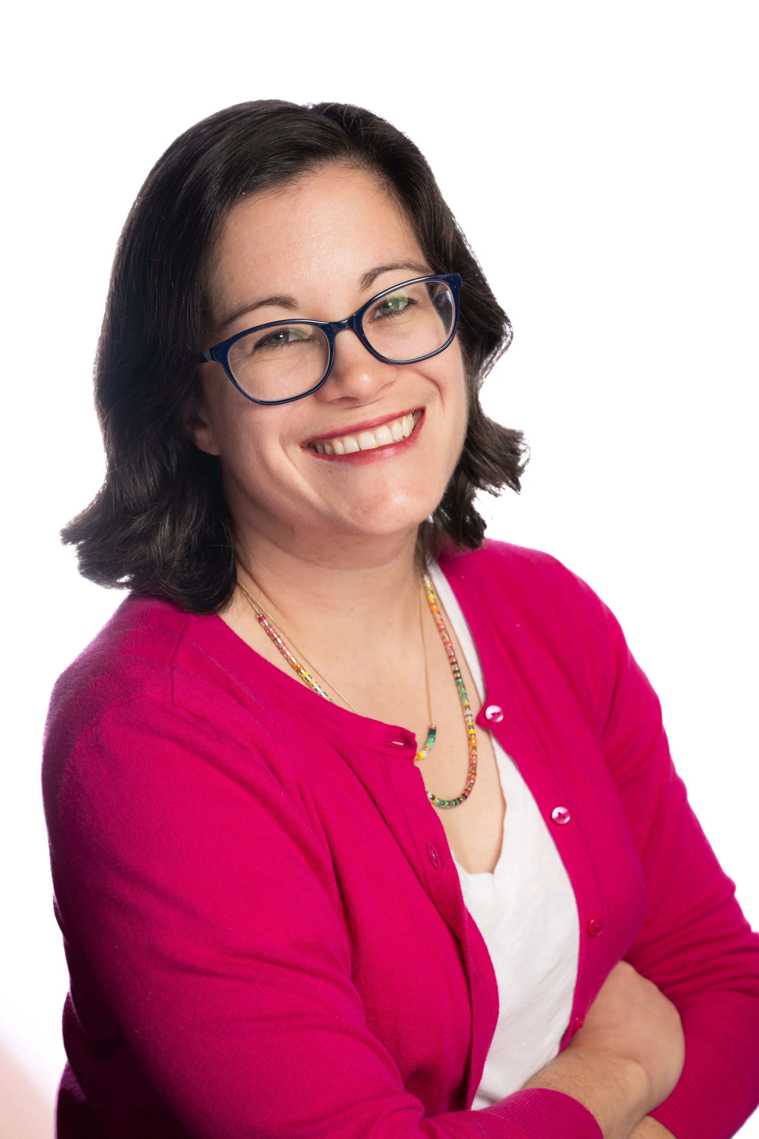 A woman with black hair, wearing glasses, a white shirt, and a pink cardigan, smiling with arms crossed against a white background.